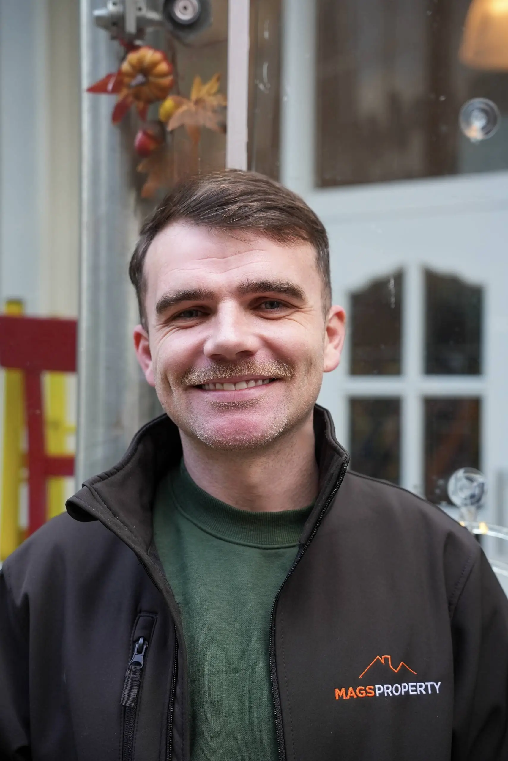 Man smiling at camera wearing black jacket with orange "Mags Property" logo over green jumper, standing in front of a building entrance