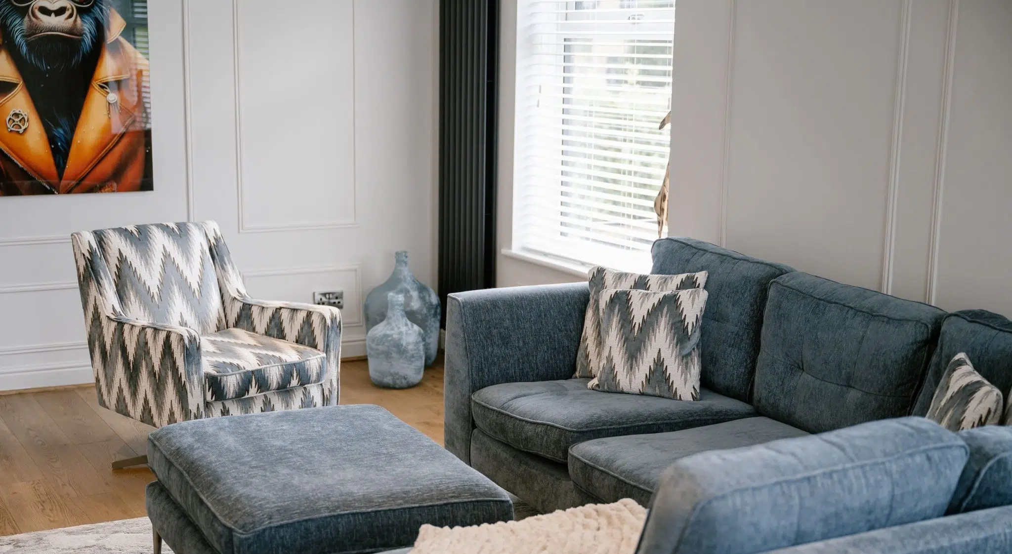 Living room with blue-grey sofa, patterned armchair, grey ottoman, and framed artwork of a chimpanzee on the wall