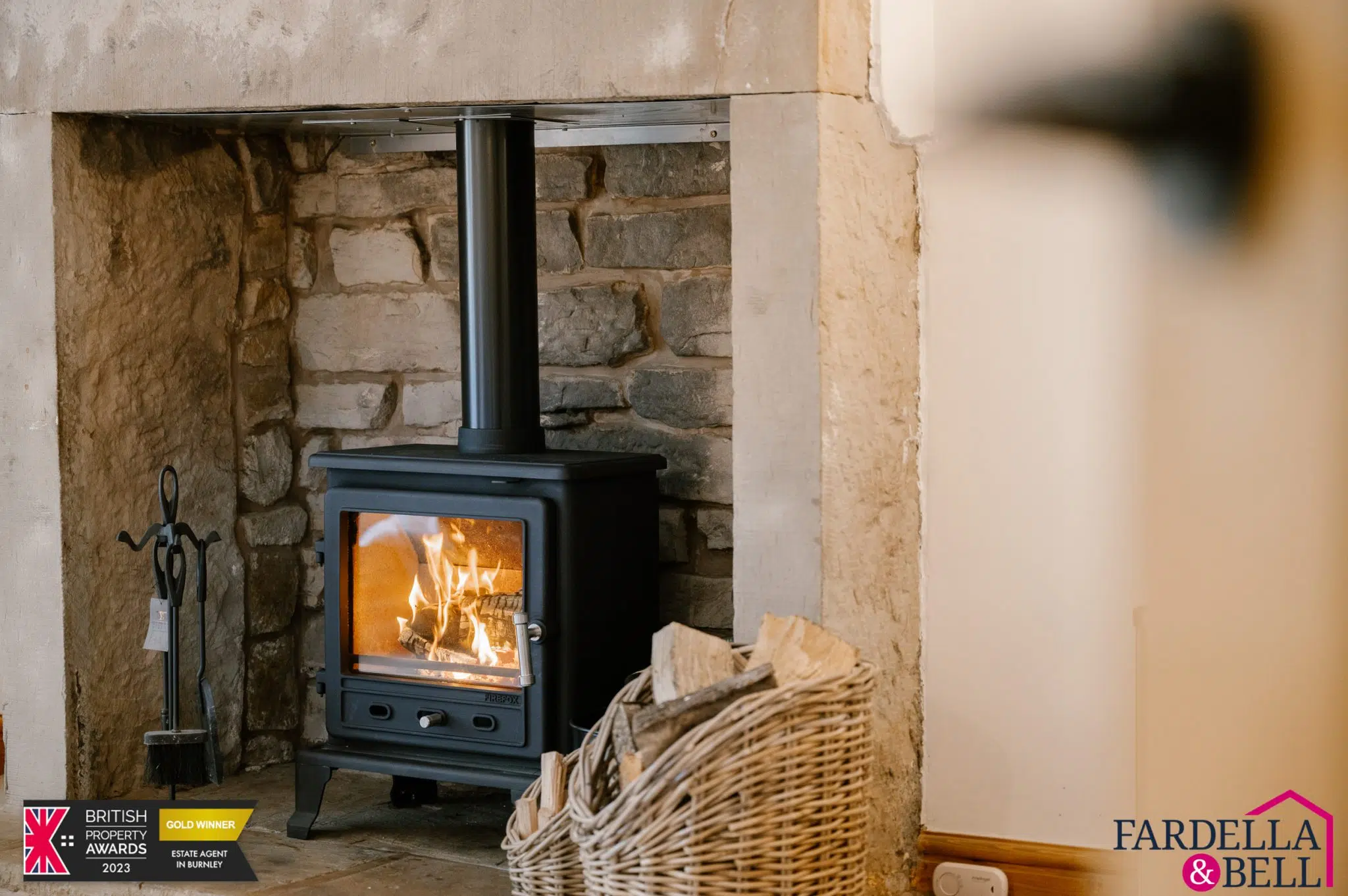 Black wood-burning stove with lit fire set into a stone fireplace, with a wicker basket of logs beside it and fireplace tools on the left