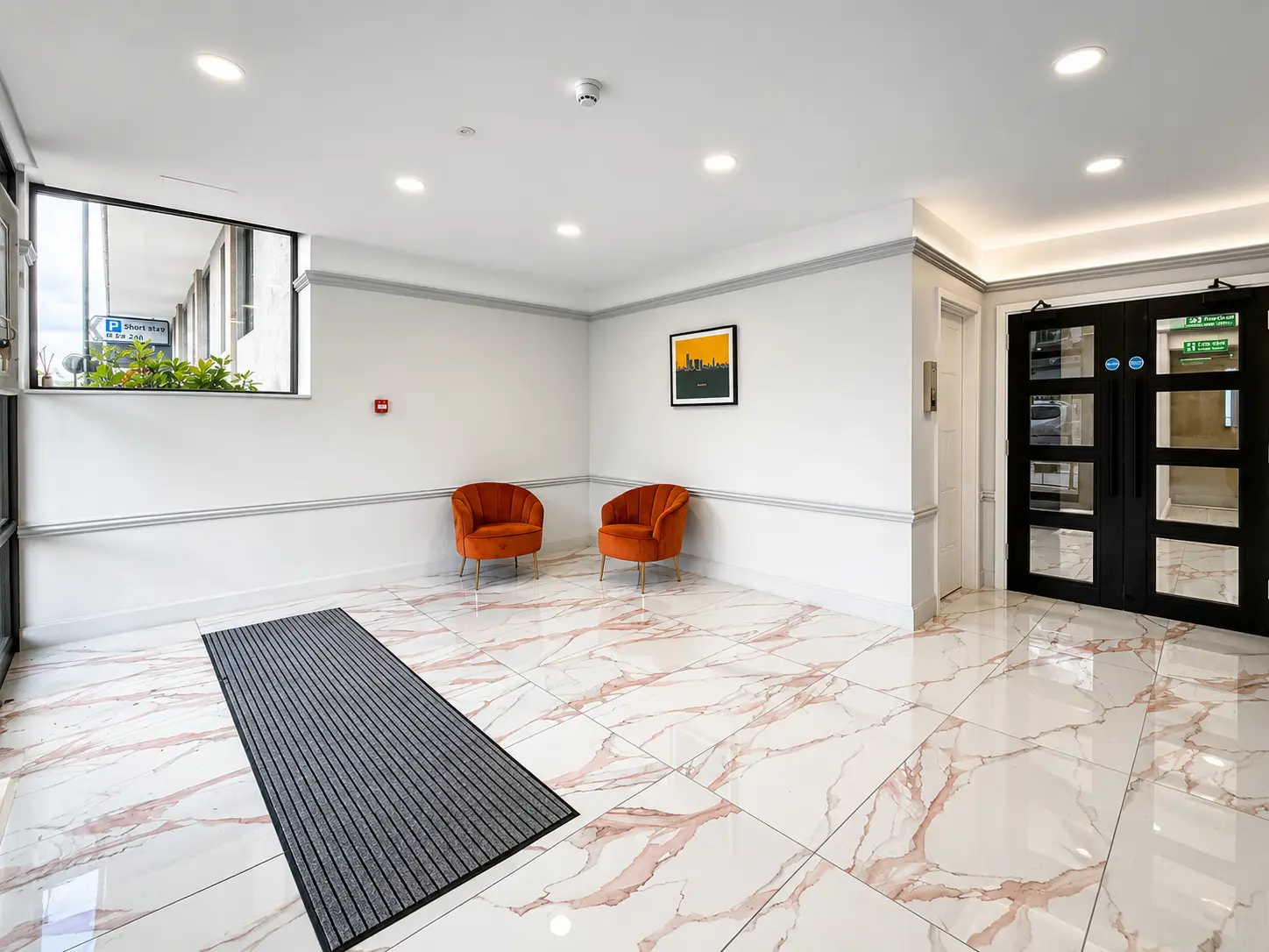 Modern building lobby with marble flooring, two orange armchairs, recessed lighting, and black double doors with glass panels on the right