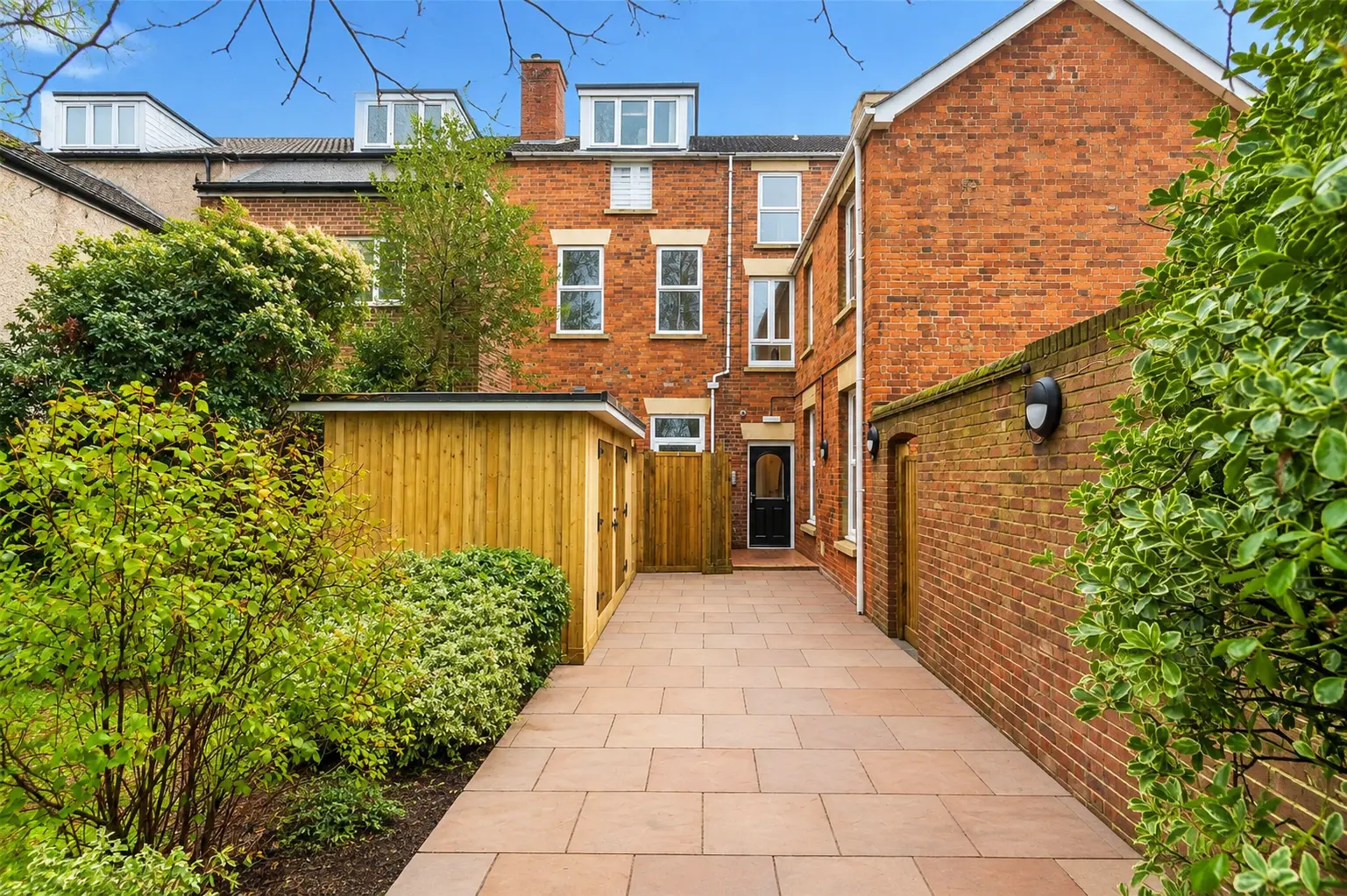 Red brick terraced house with paved courtyard, yellow wooden garage doors, and surrounding hedges and greenery