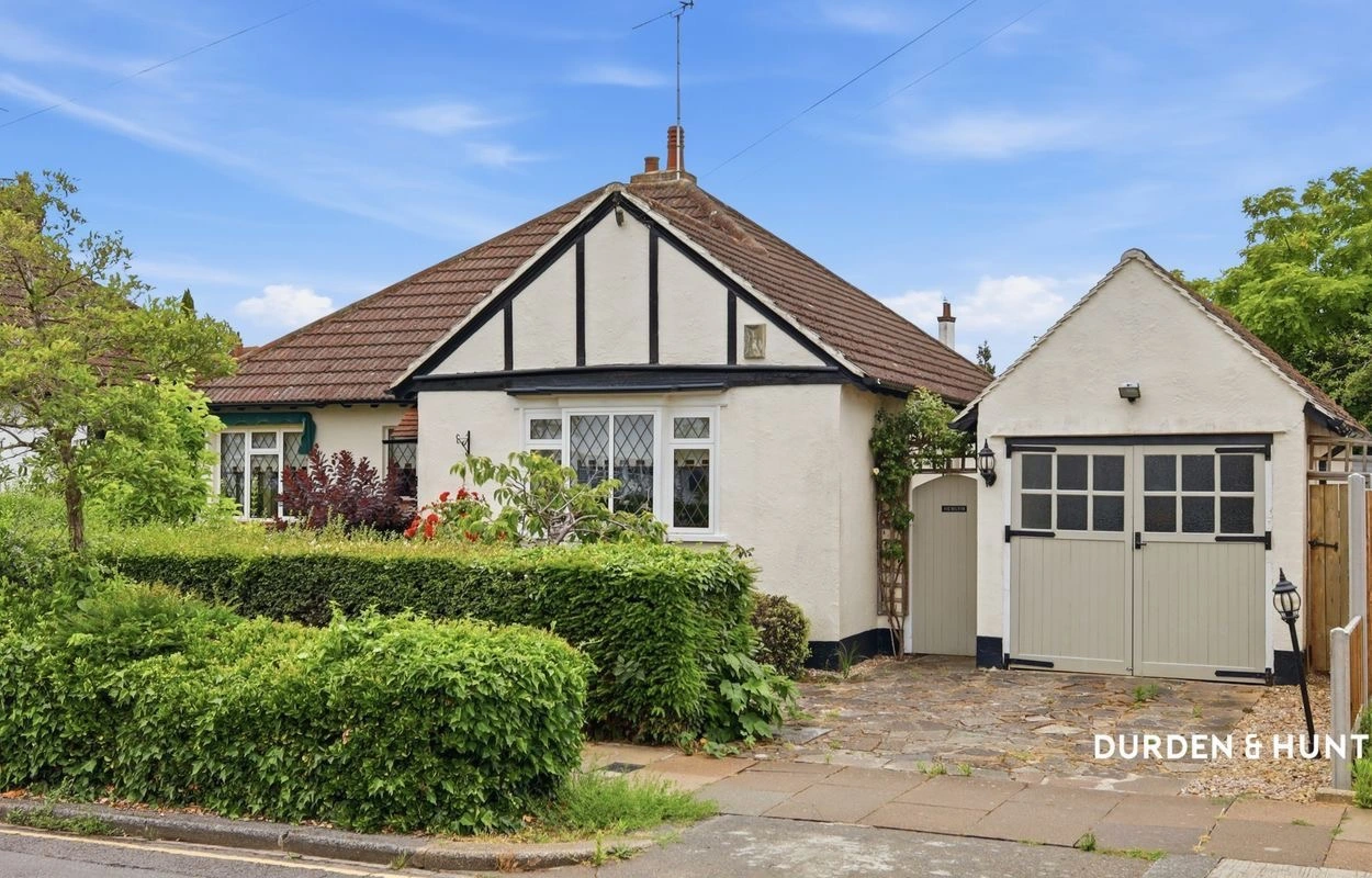White rendered bungalow with brown tiled roof and black timber framing, grey garage, mature hedging and landscaped front garden