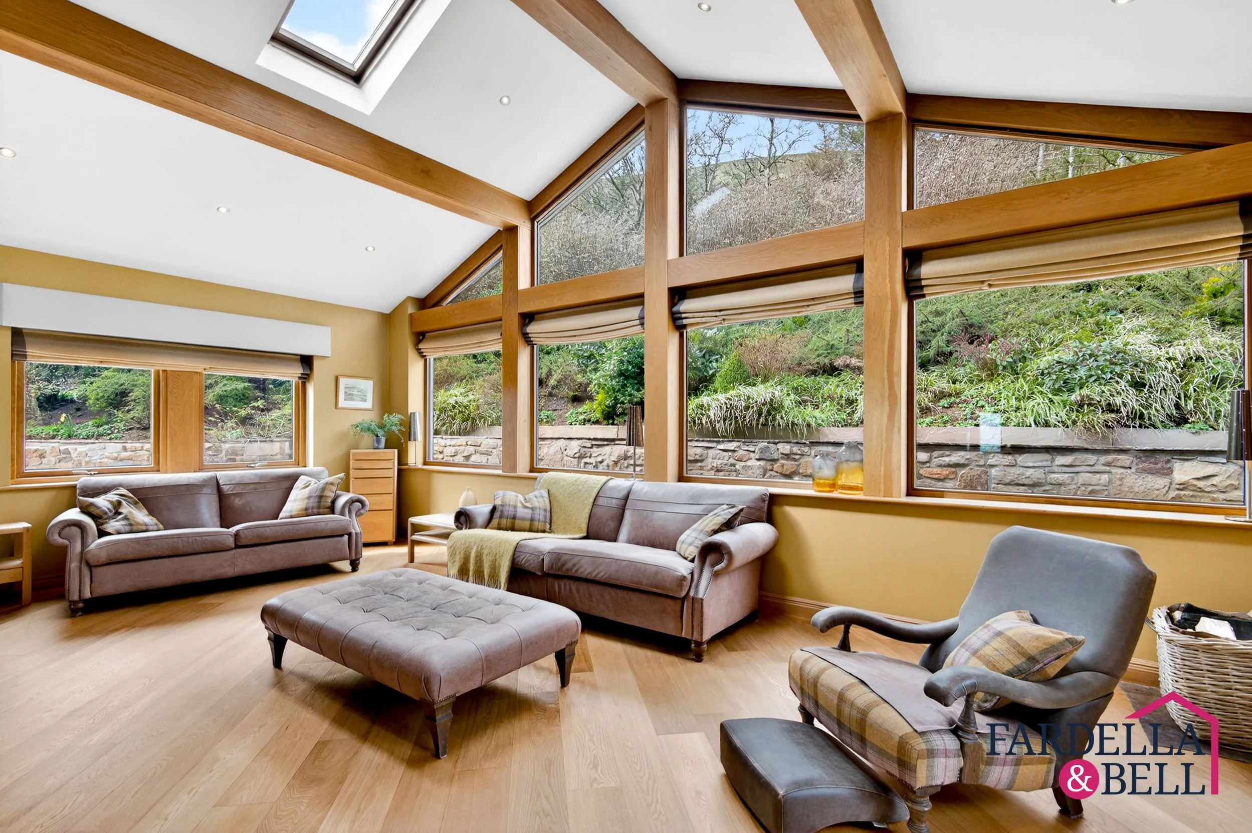Modern conservatory with wooden beams, skylights, and large windows overlooking a garden. Brown leather sofas and armchairs with cushions, a quilted ottoman, and wooden flooring. Yellow-painted walls and stone garden wall visible through windows.