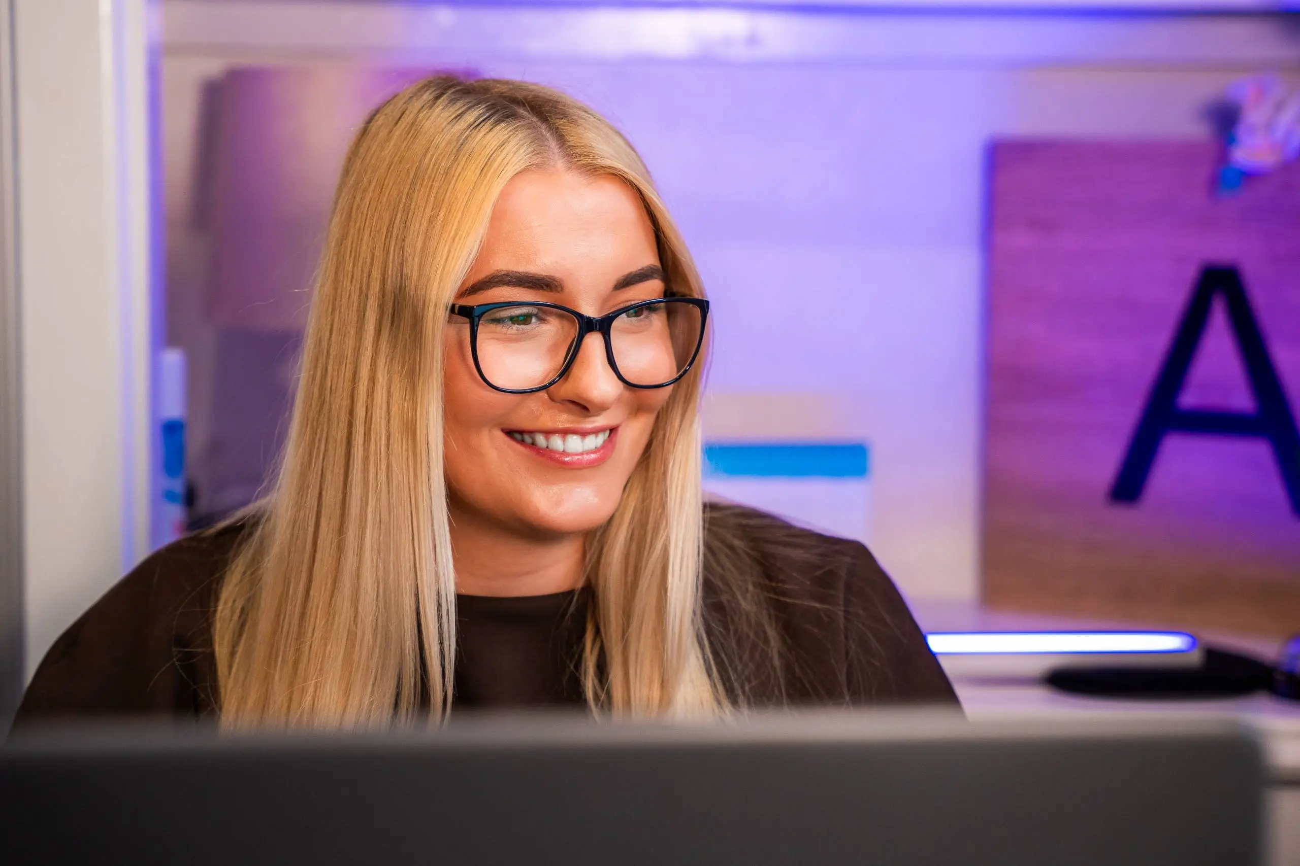 Person with blonde hair and glasses smiling at a desk with purple-lit office interior in background