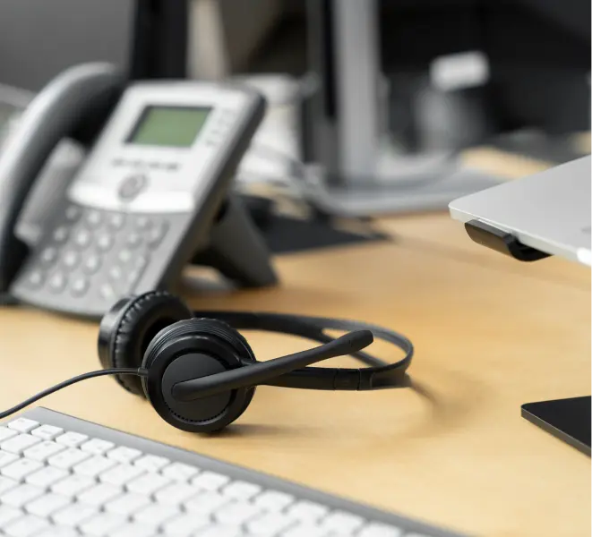 Black headset with microphone on wooden desk next to white keyboard and office telephone