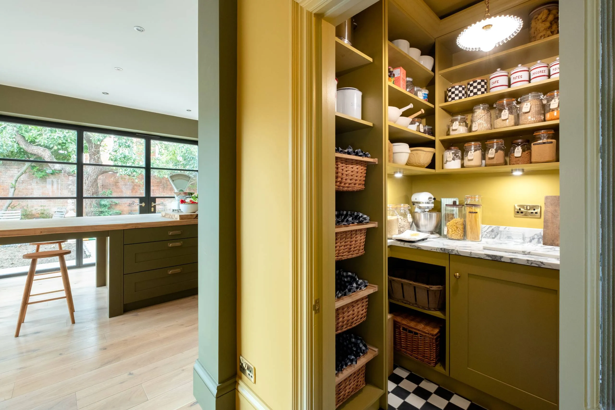 Mustard yellow butler's pantry with open shelving, wicker baskets and marble worktop, leading to a green kitchen with checkerboard floor