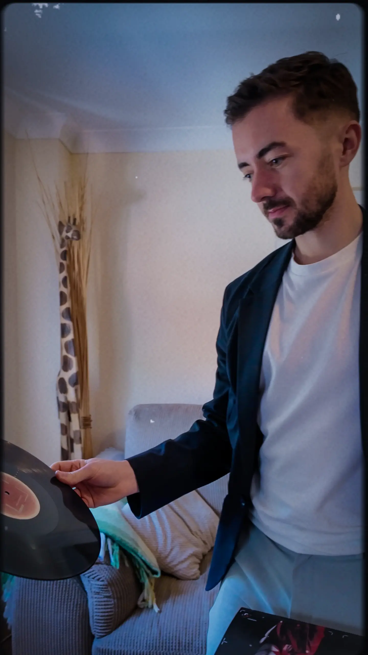 Man in dark blazer and white shirt holding a tablet, standing in a bright interior space with dried grasses in a vase