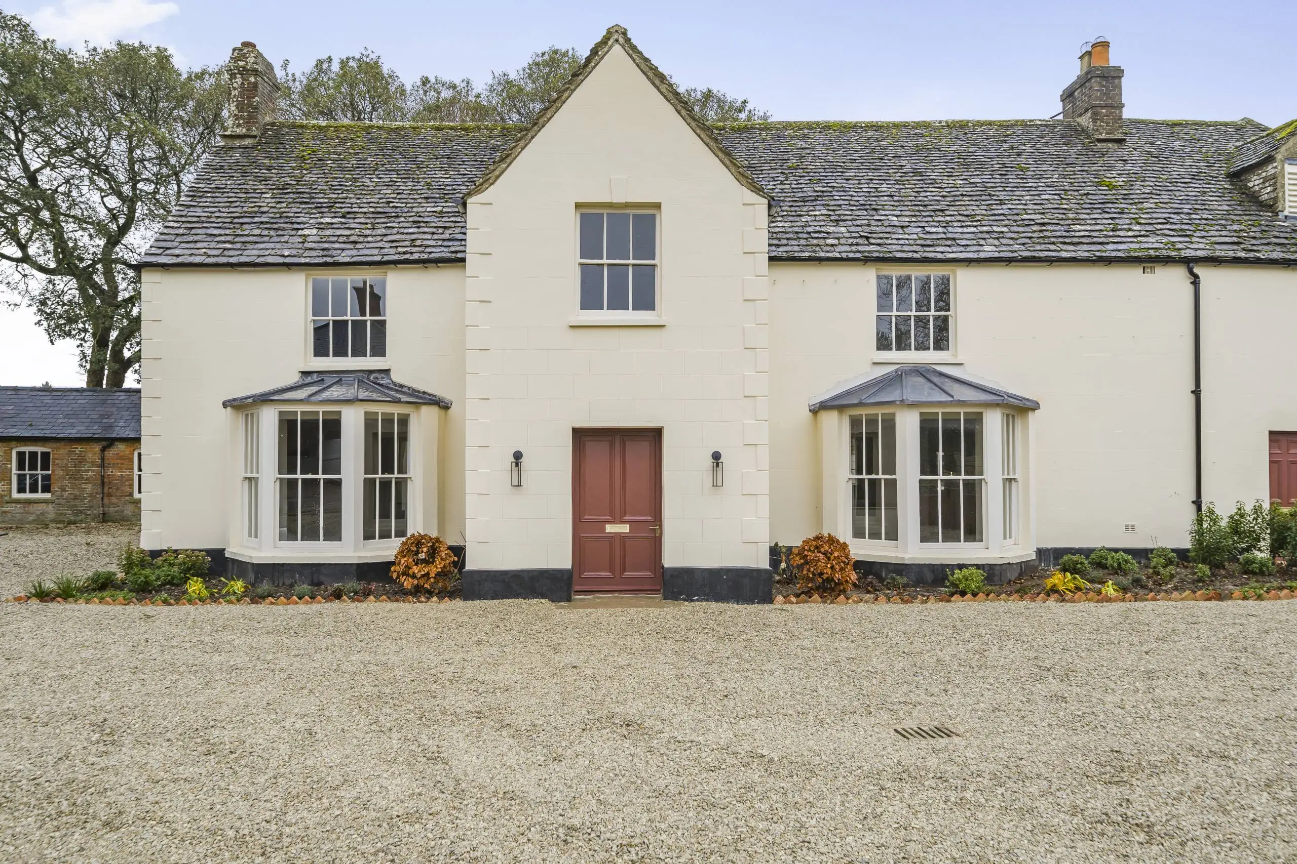 Cream-coloured country house with slate roof, central red door, symmetrical bay windows with pitched roofs, and gravel driveway