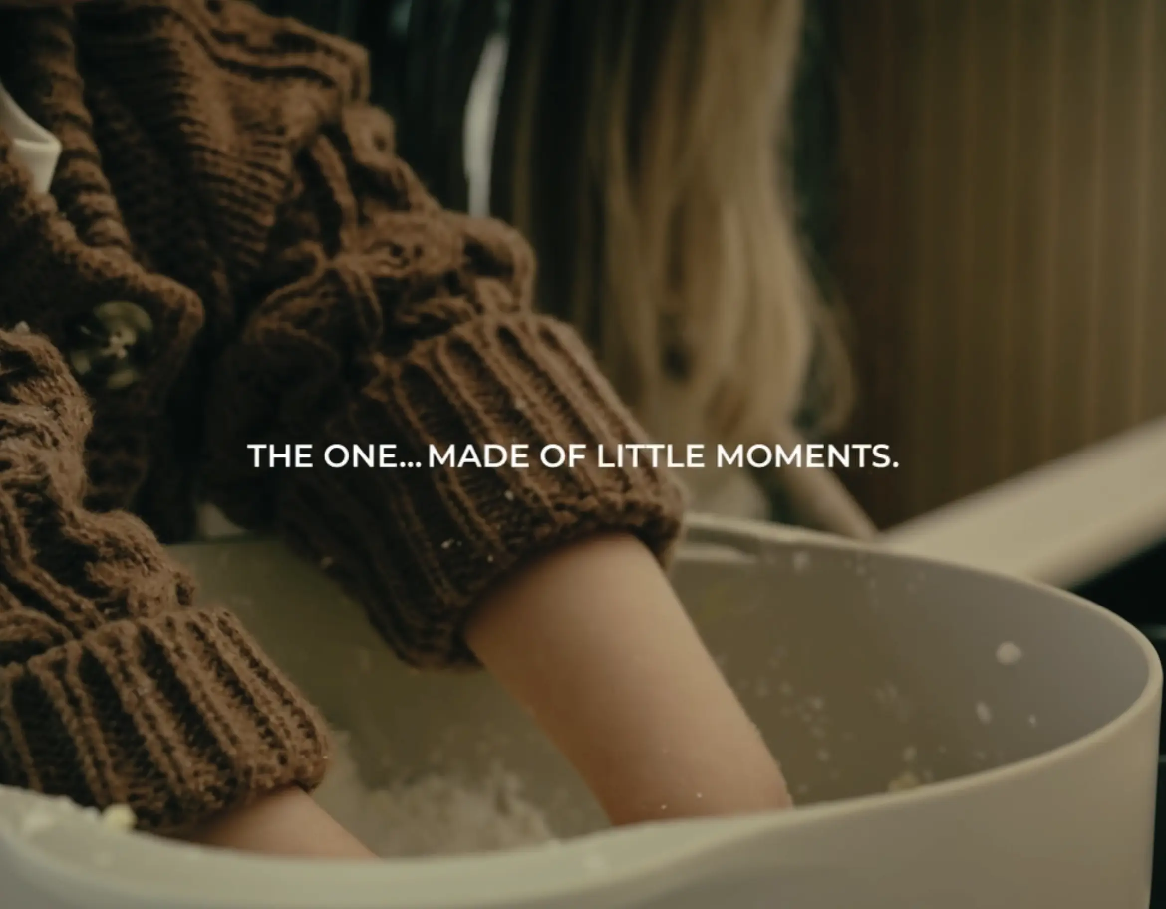 Person wearing brown knitted gloves dipping hands into a white bowl of water, with text overlay reading 'THE ONE...MADE OF LITTLE MOMENTS.'