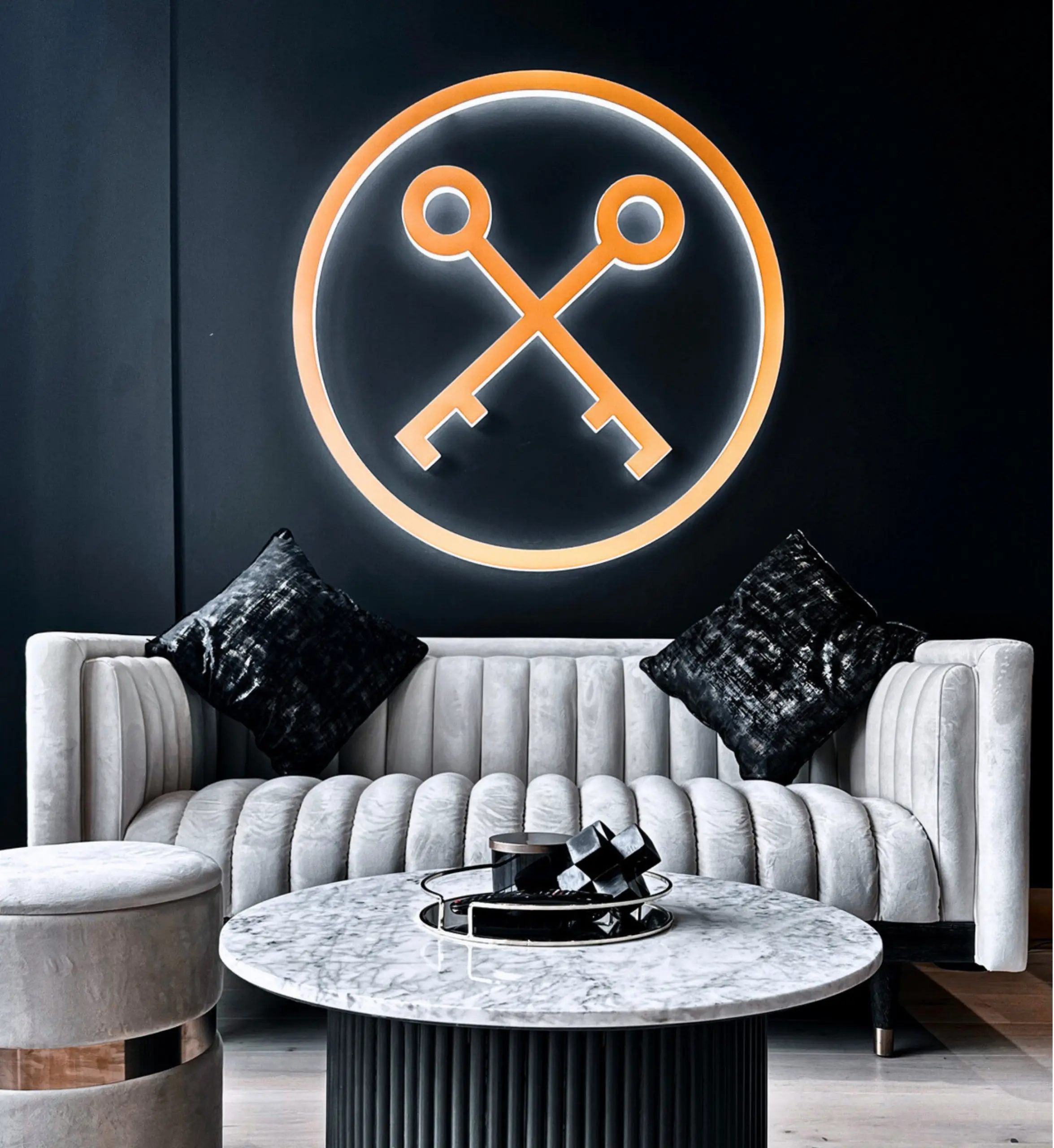 Modern interior with cream ribbed sofa, black cushions, marble-top table, and orange neon crossed-keys symbol on dark wall