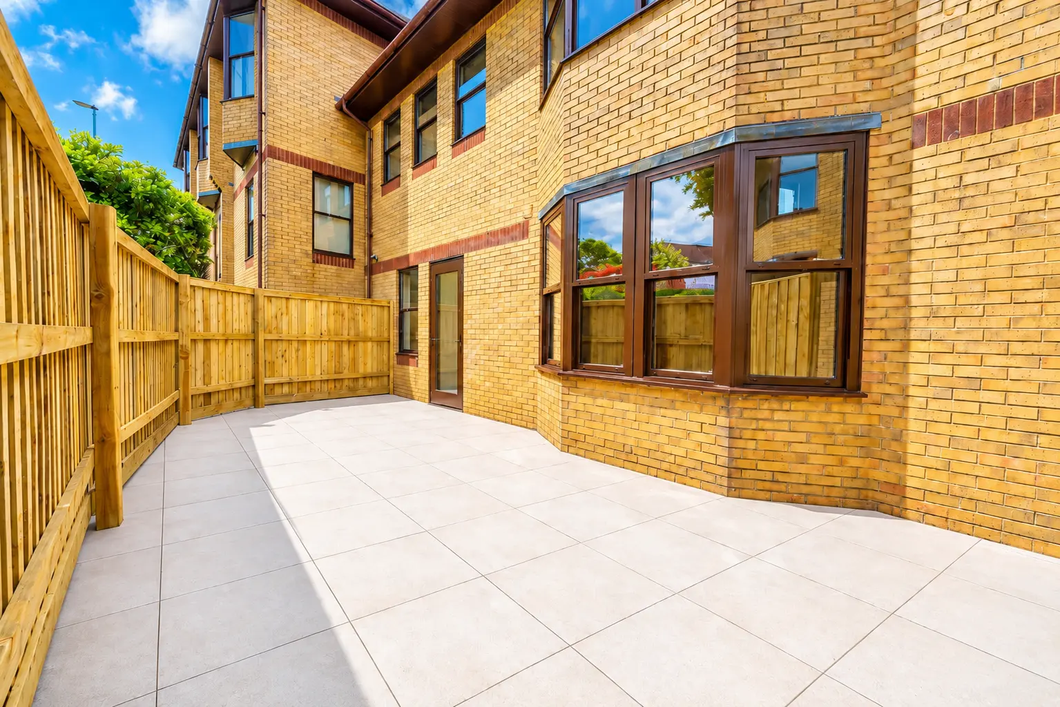 Paved patio area with wooden fence, brick building with brown-framed windows and glass doors