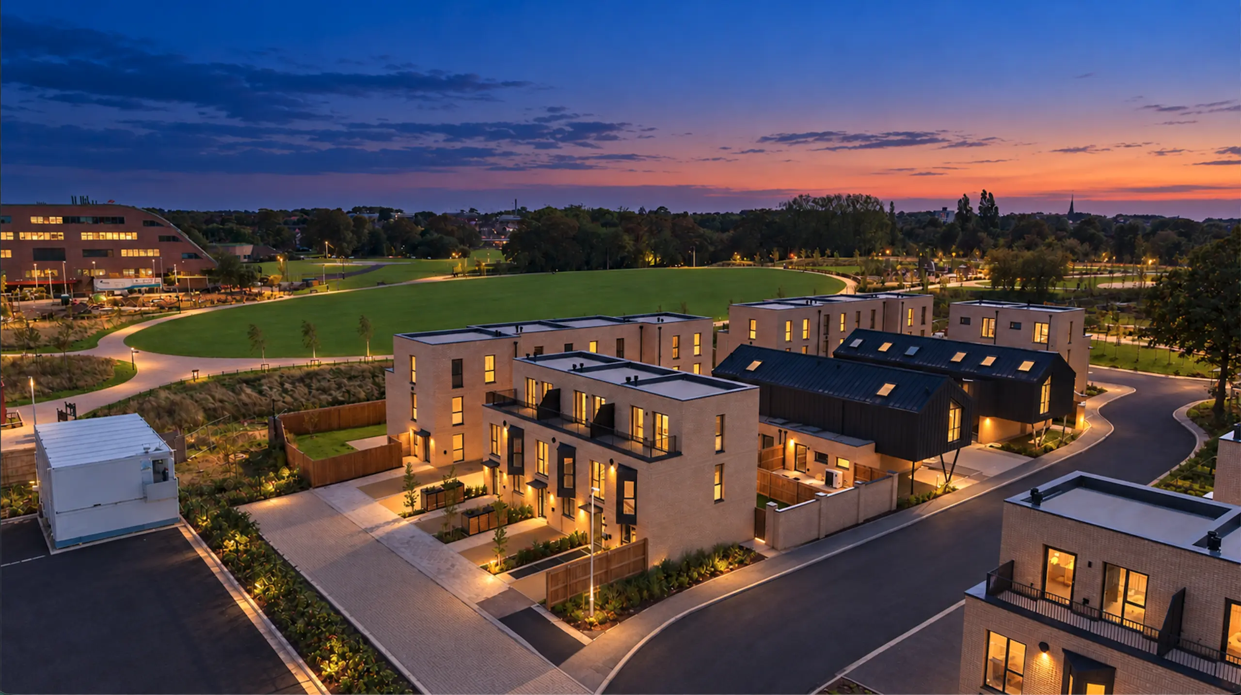 New residential development at dusk with modern brick houses, lit windows, paved streets, landscaped grounds and green space beyond
