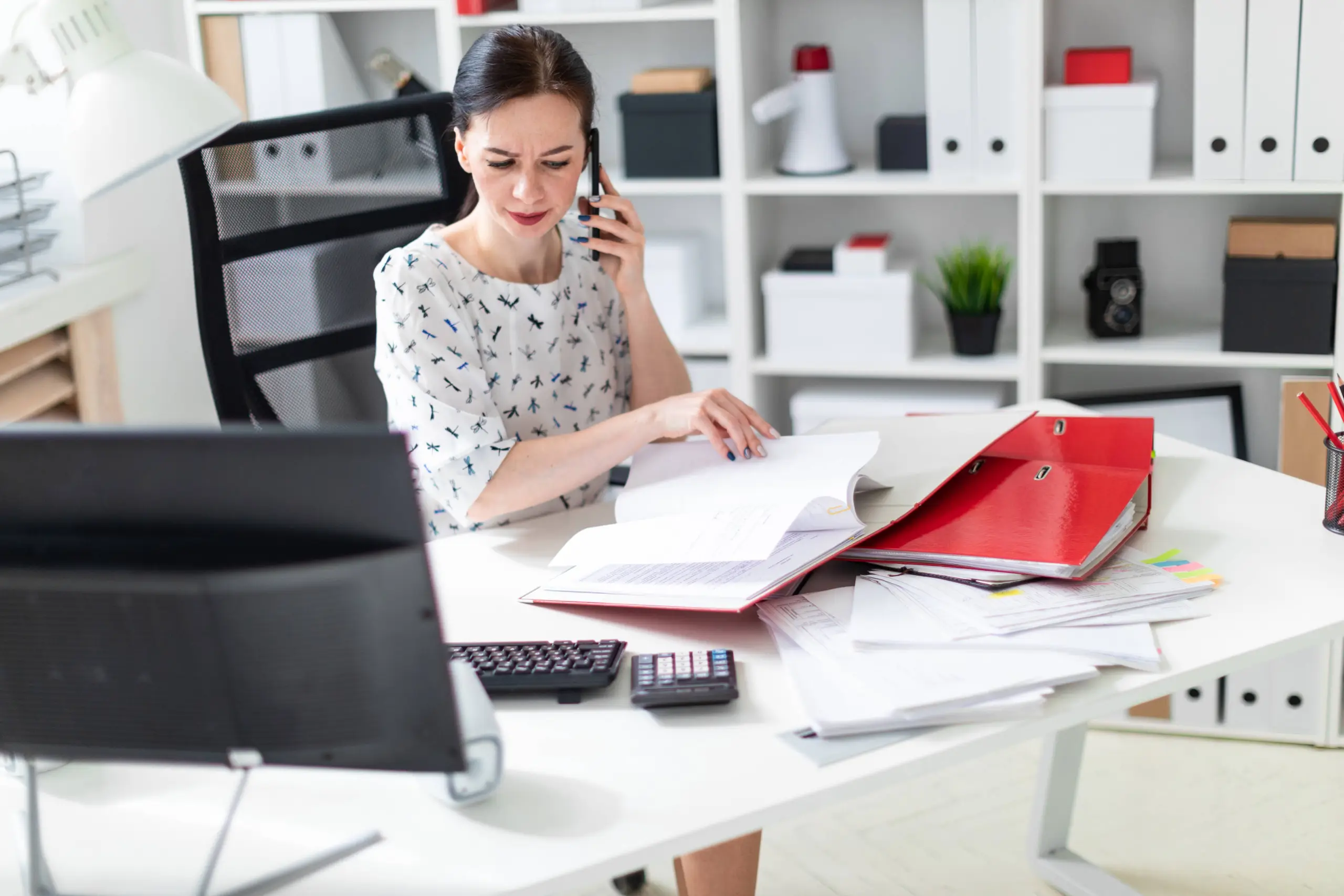 Woman at desk in office talking on phone while reviewing documents, with laptop, red folders, and calculator nearby