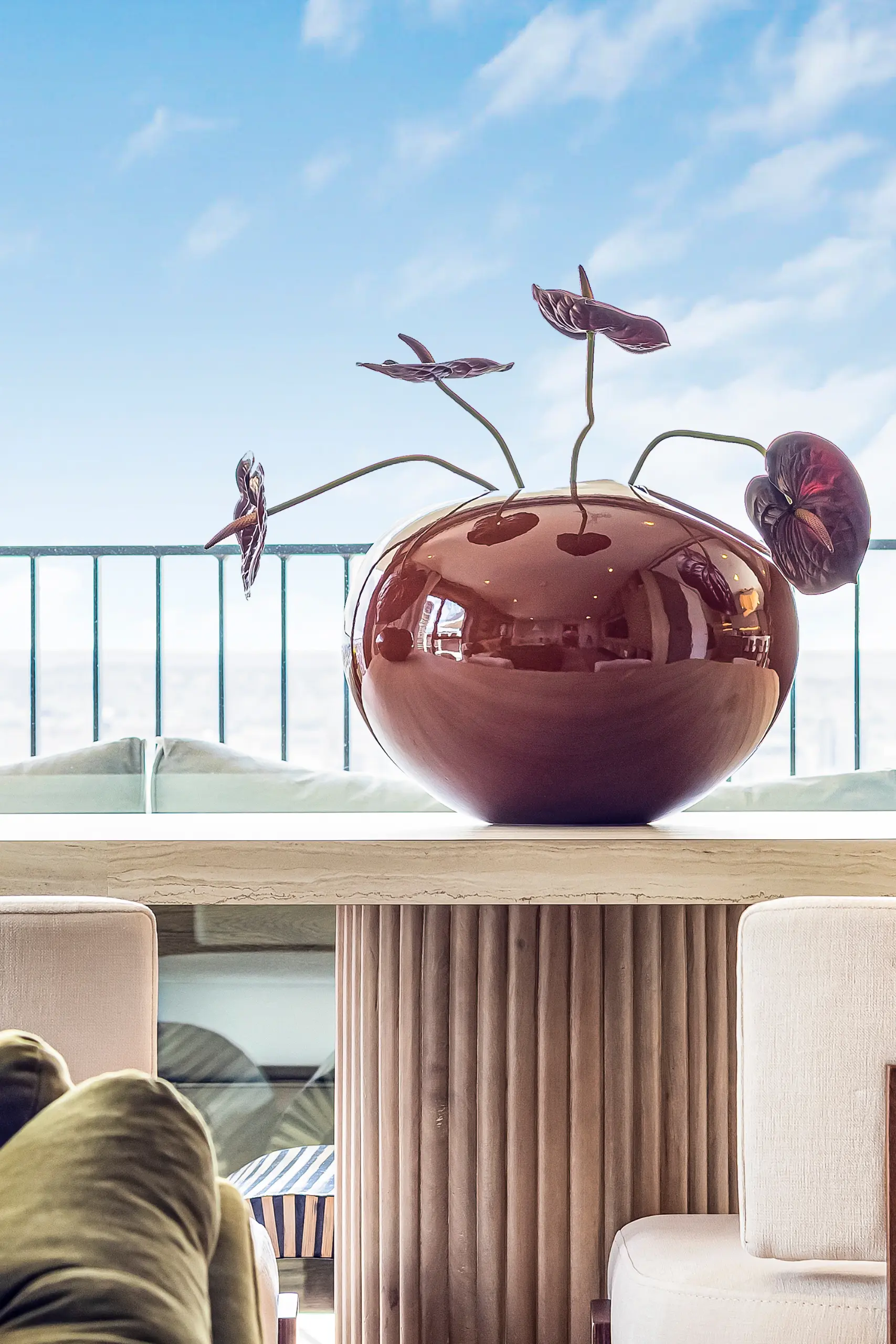 Two burgundy ceramic spherical planters with wilting flowers on a wooden windowsill, overlooking a balcony with metal railings and sky