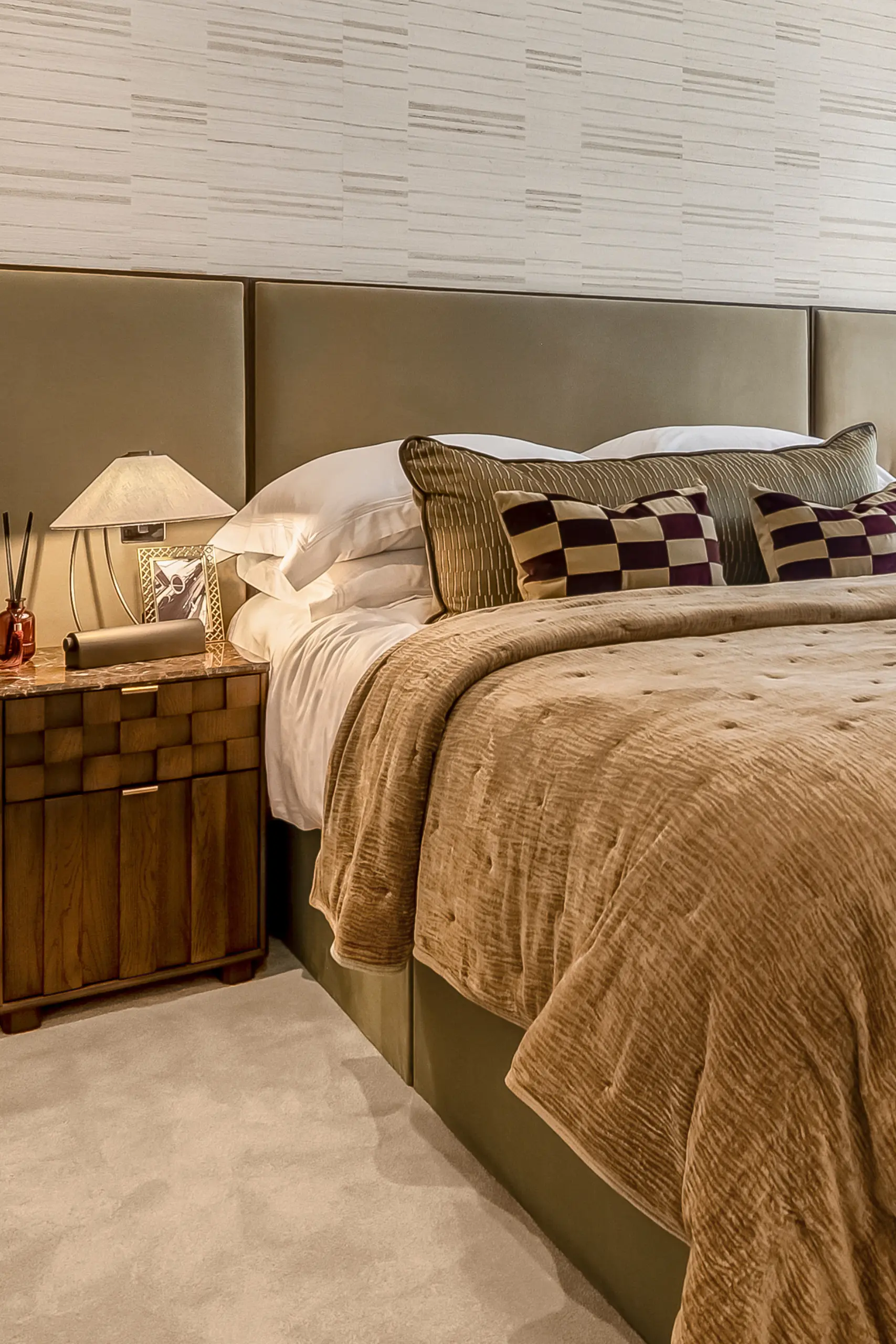 Bedroom with upholstered headboard, wooden bedside table with lamp, and neutral-toned bedding and furnishings