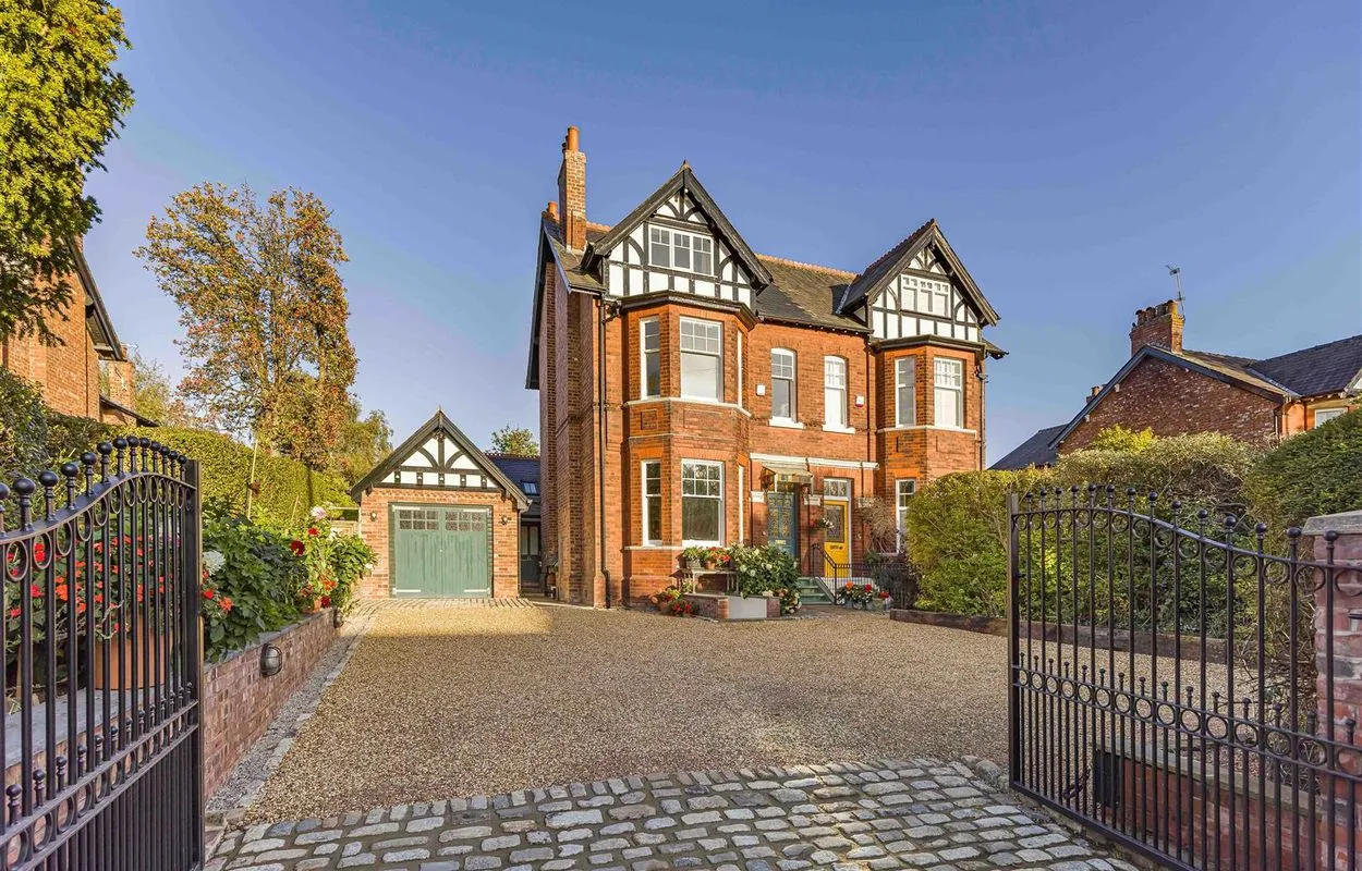 Victorian red brick house with black and white half-timbered gables, featuring a green garage door, gravel driveway, and black iron gates