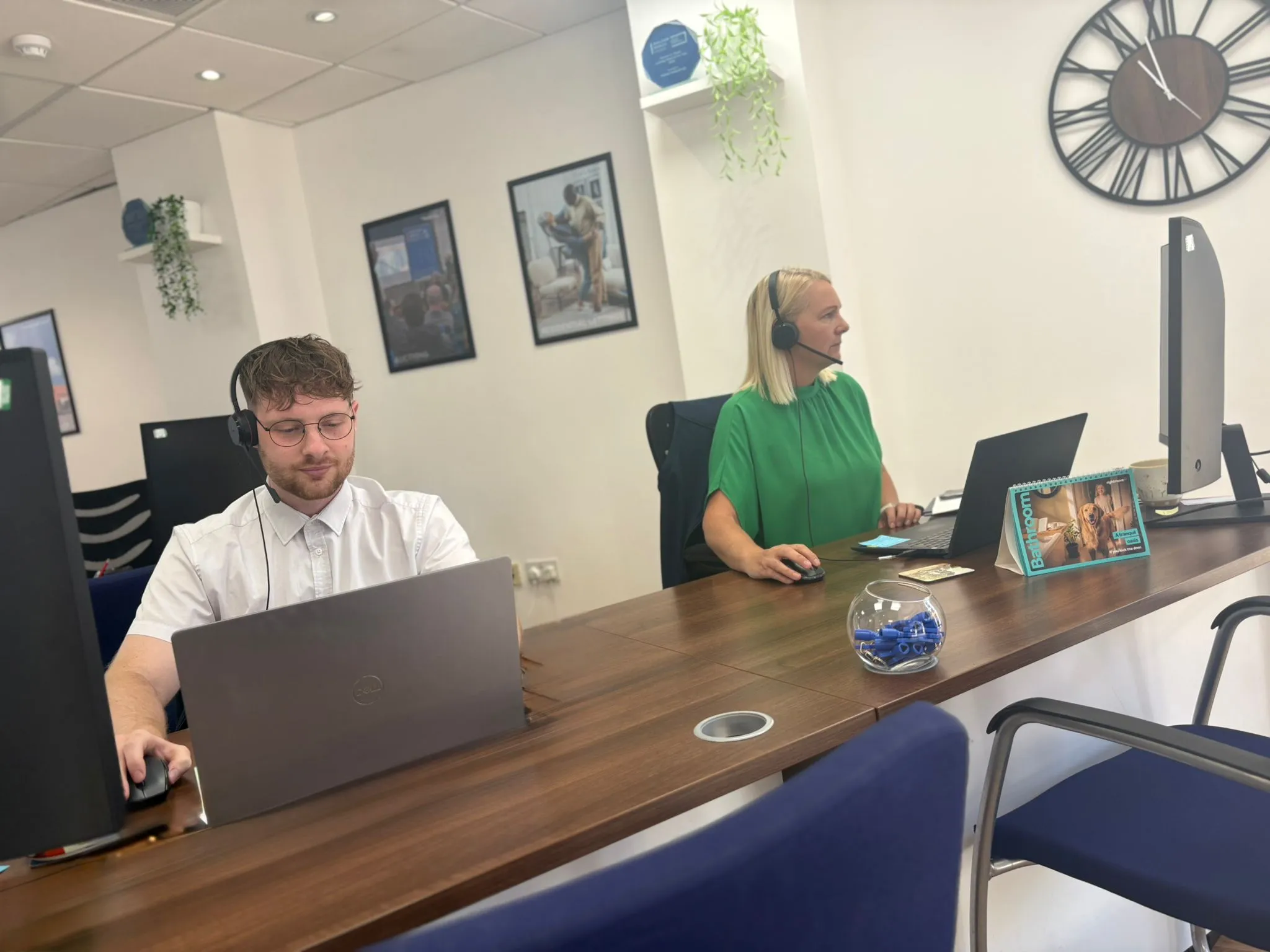 Two people working at desks in an office. Man on left wearing white shirt and headphones, working on laptop. Woman on right wearing green shirt and headphones, working on laptop. Wooden desk with decorative items including a blue globe and framed photos on walls behind.