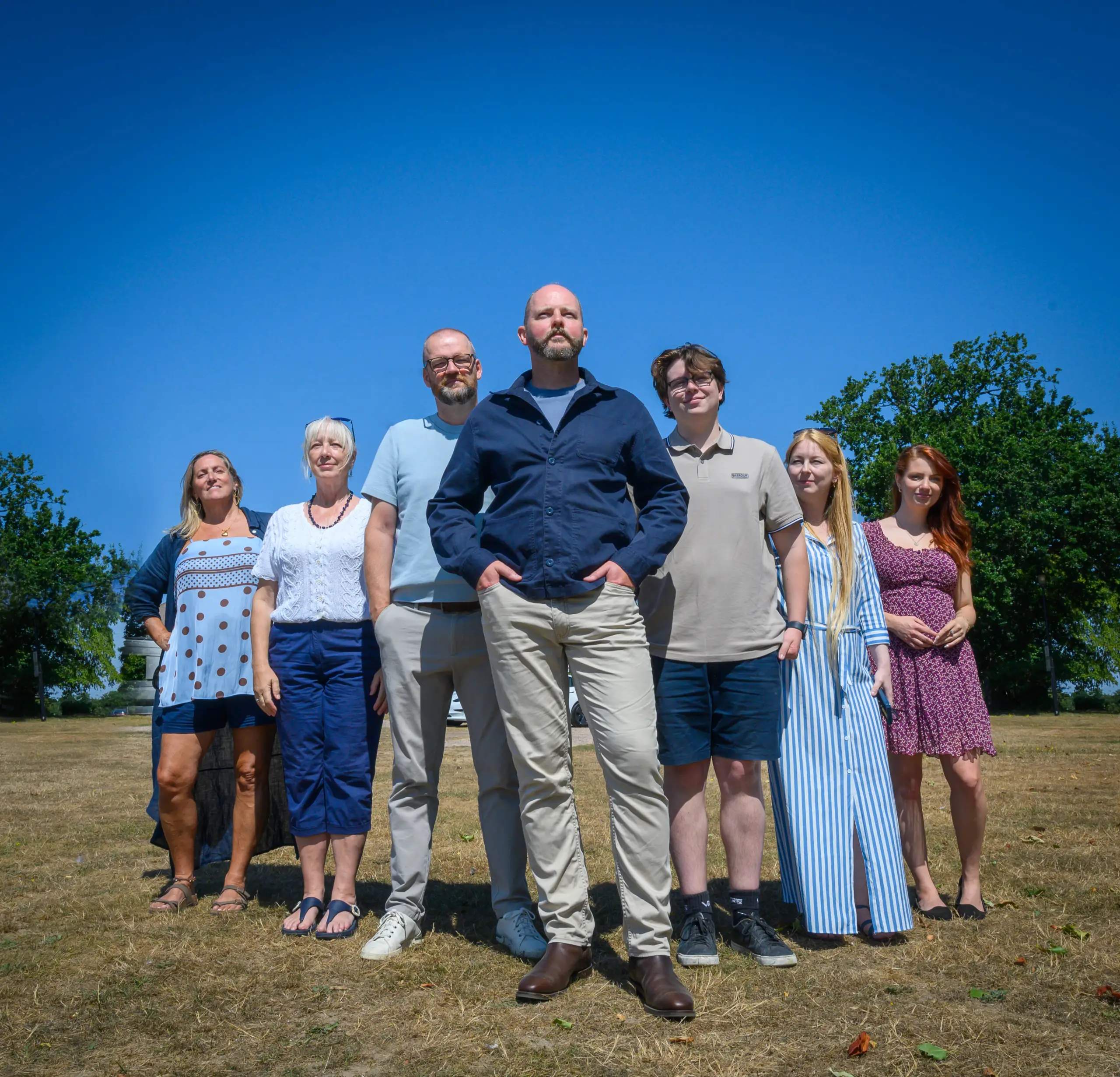 Seven people standing in a line outdoors on a grass field with trees and clear blue sky in the background