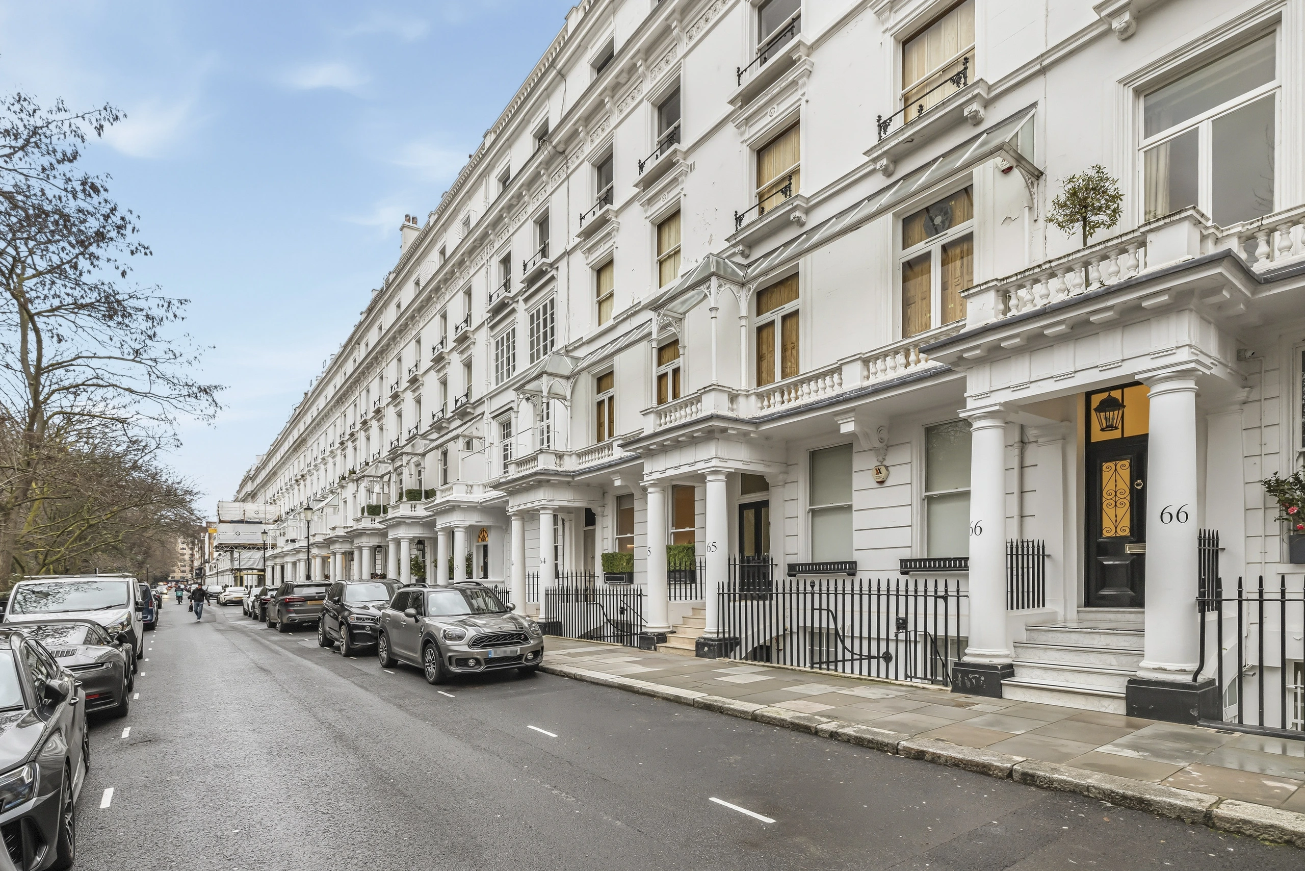 White stucco period building with columned portico, numbered 65 and 66, on a tree-lined London street with parked cars