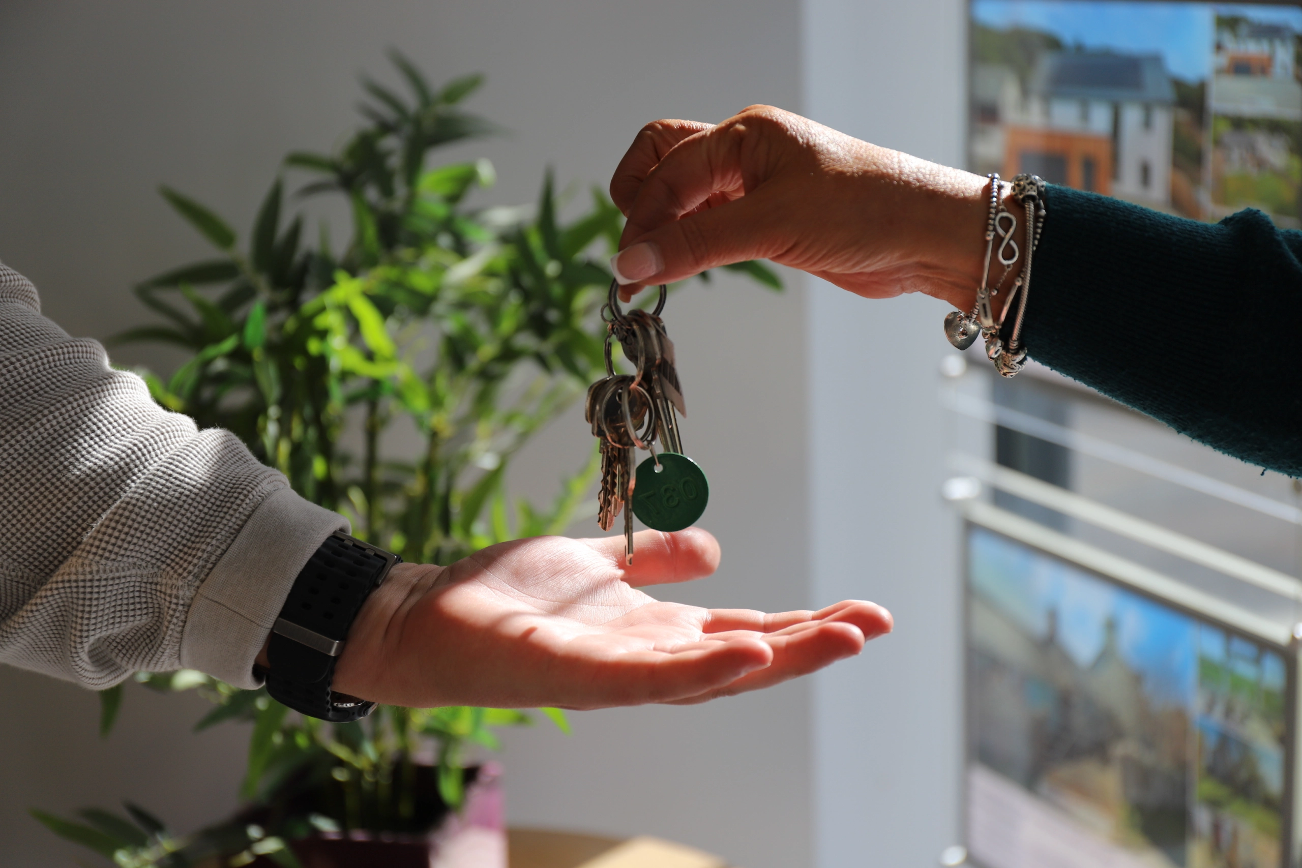 Two hands holding a set of keys with a green circular keyring, with potted plants and residential buildings visible in the background