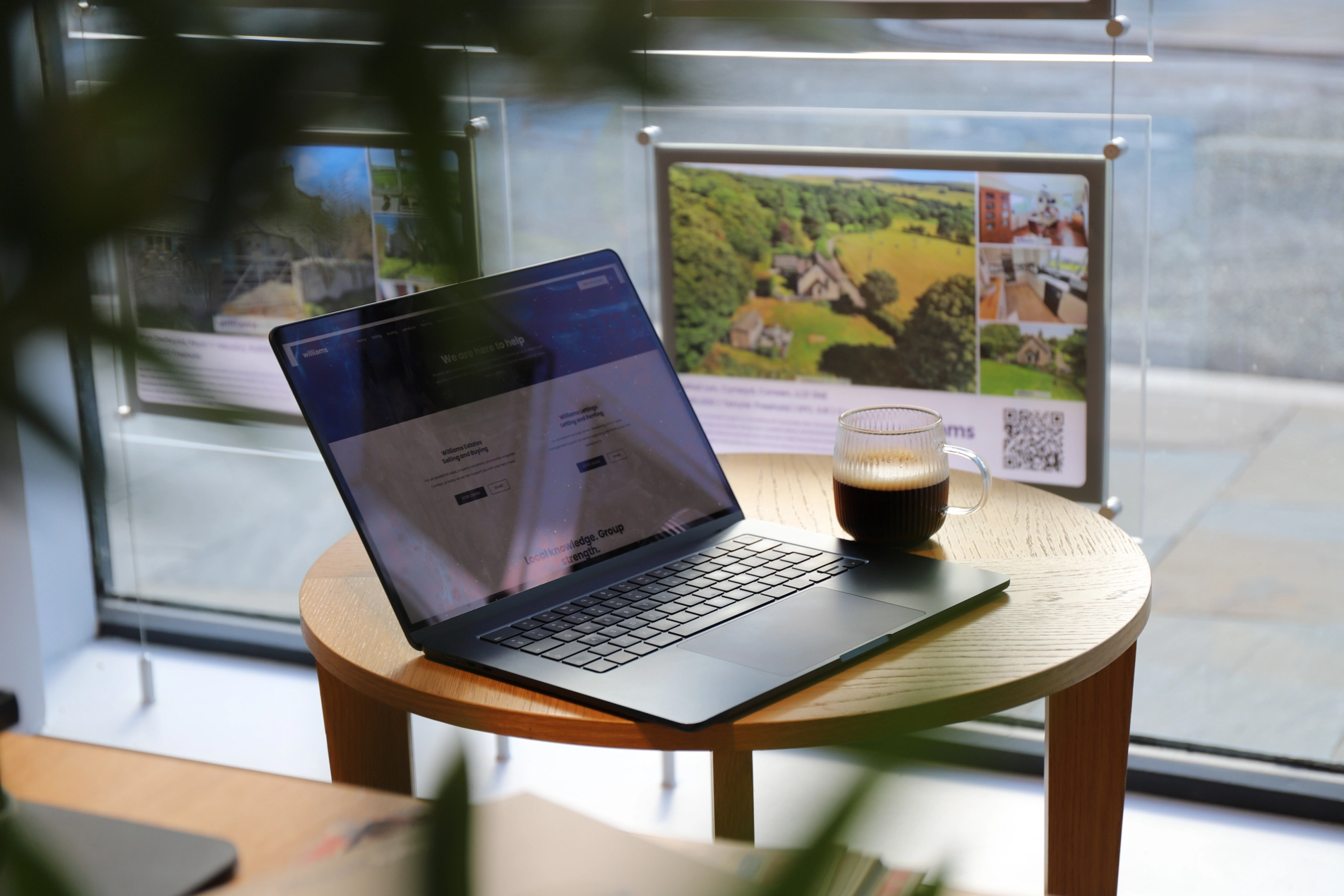 Laptop displaying a website on a wooden table with a coffee mug, framed property photographs visible in background