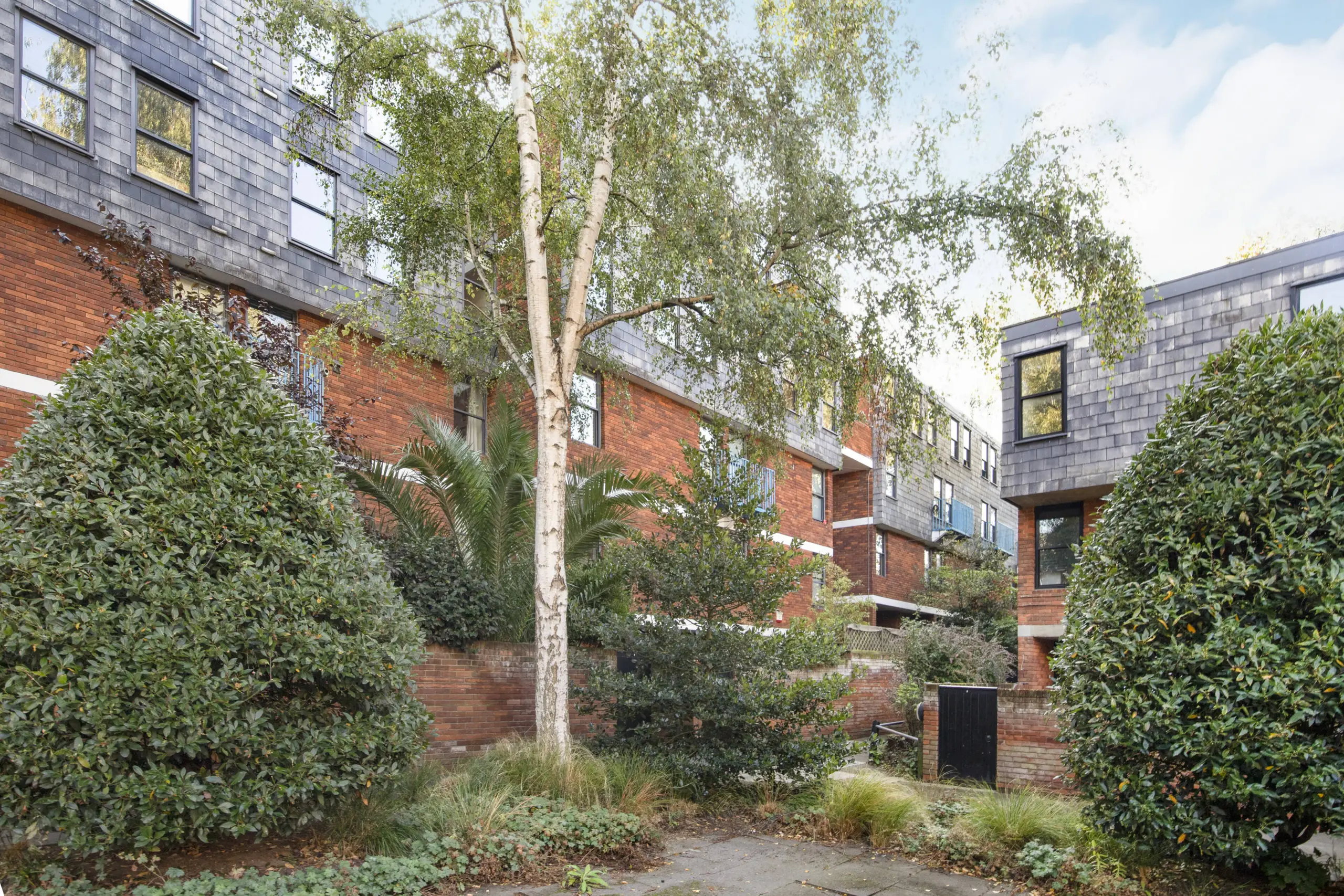Courtyard between brick and slate-clad residential buildings with mature trees, hedges and planted borders