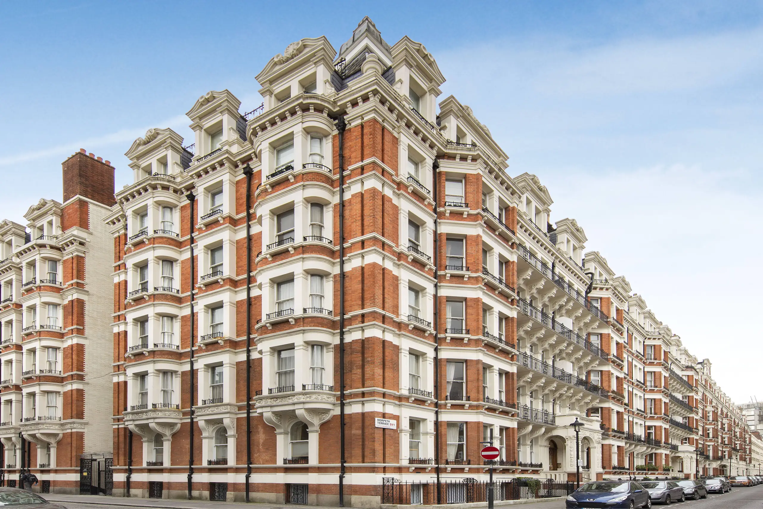 Victorian mansion block with red brick and cream stone facade, ornate cornicing and mansard roofs, parked cars at street level