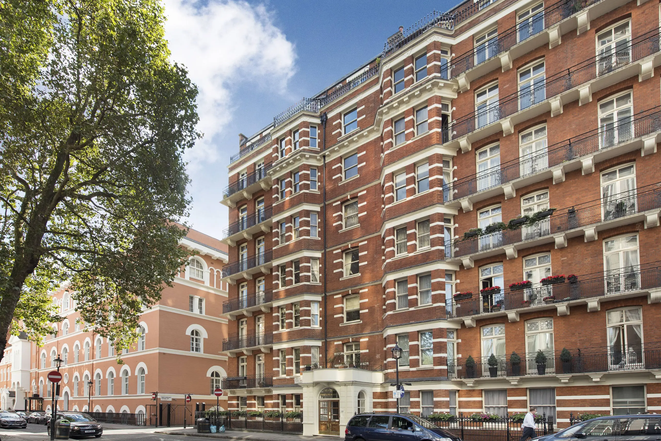 Red brick Victorian mansion building with cream stone detailing, multiple storeys and balconies, adjacent to a pink-rendered building, tree and street-level railings visible