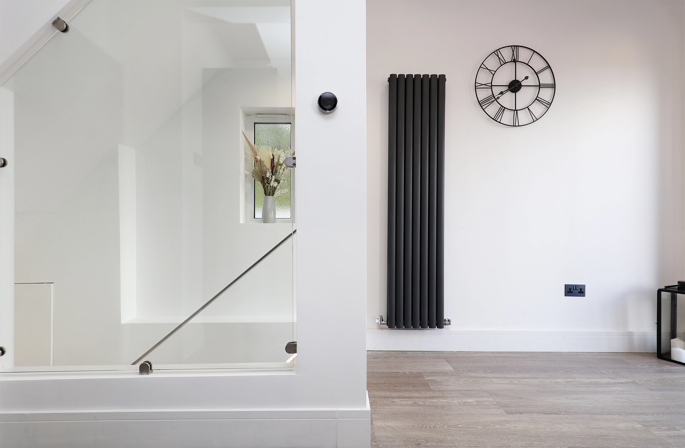 Modern interior with white walls, black vertical radiator, Roman numeral wall clock, and staircase with glass balustrade