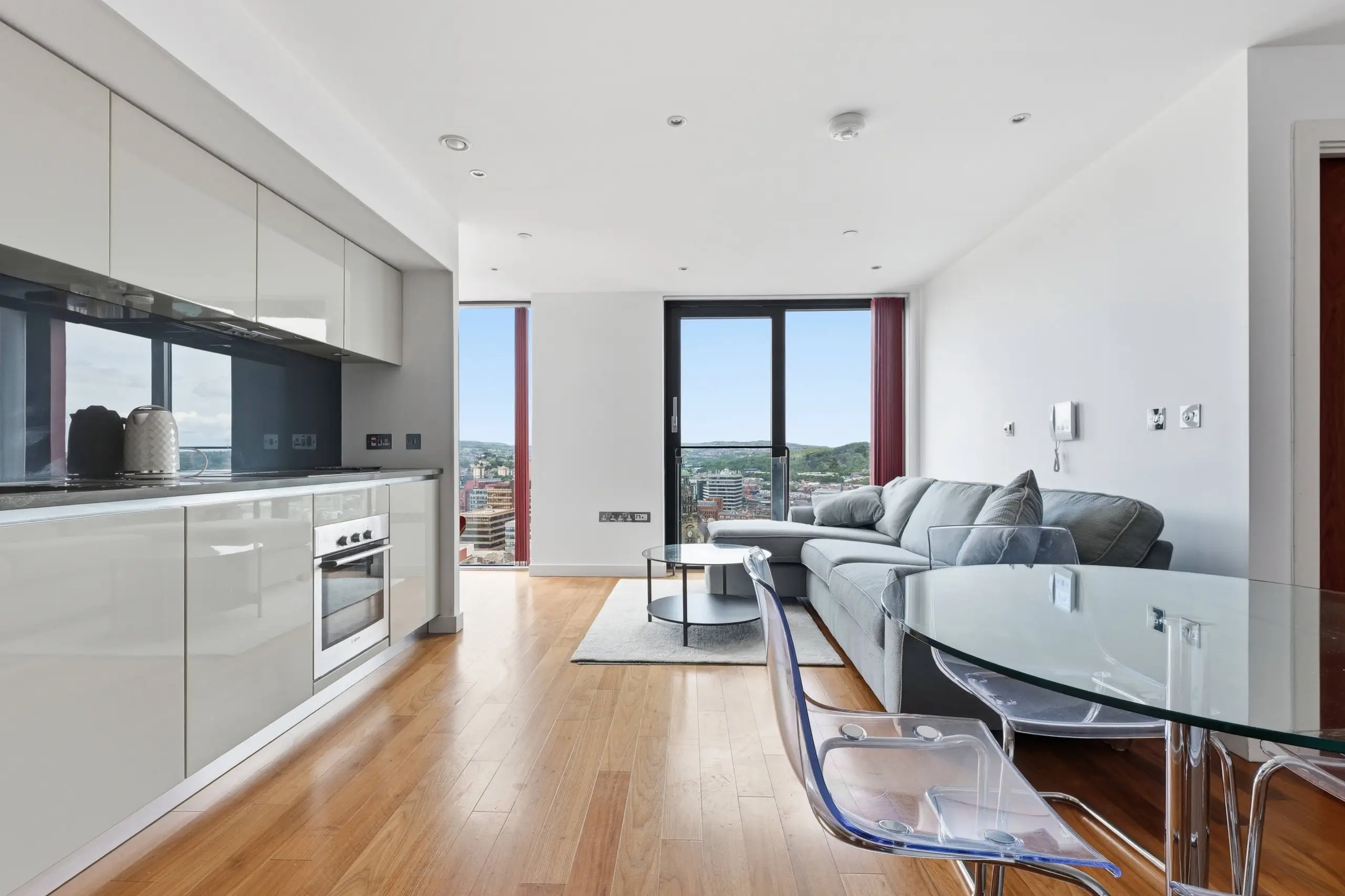 Open-plan kitchen and living area with white gloss cabinetry, black splashback, wooden flooring, grey sofa, and floor-to-ceiling windows with city views