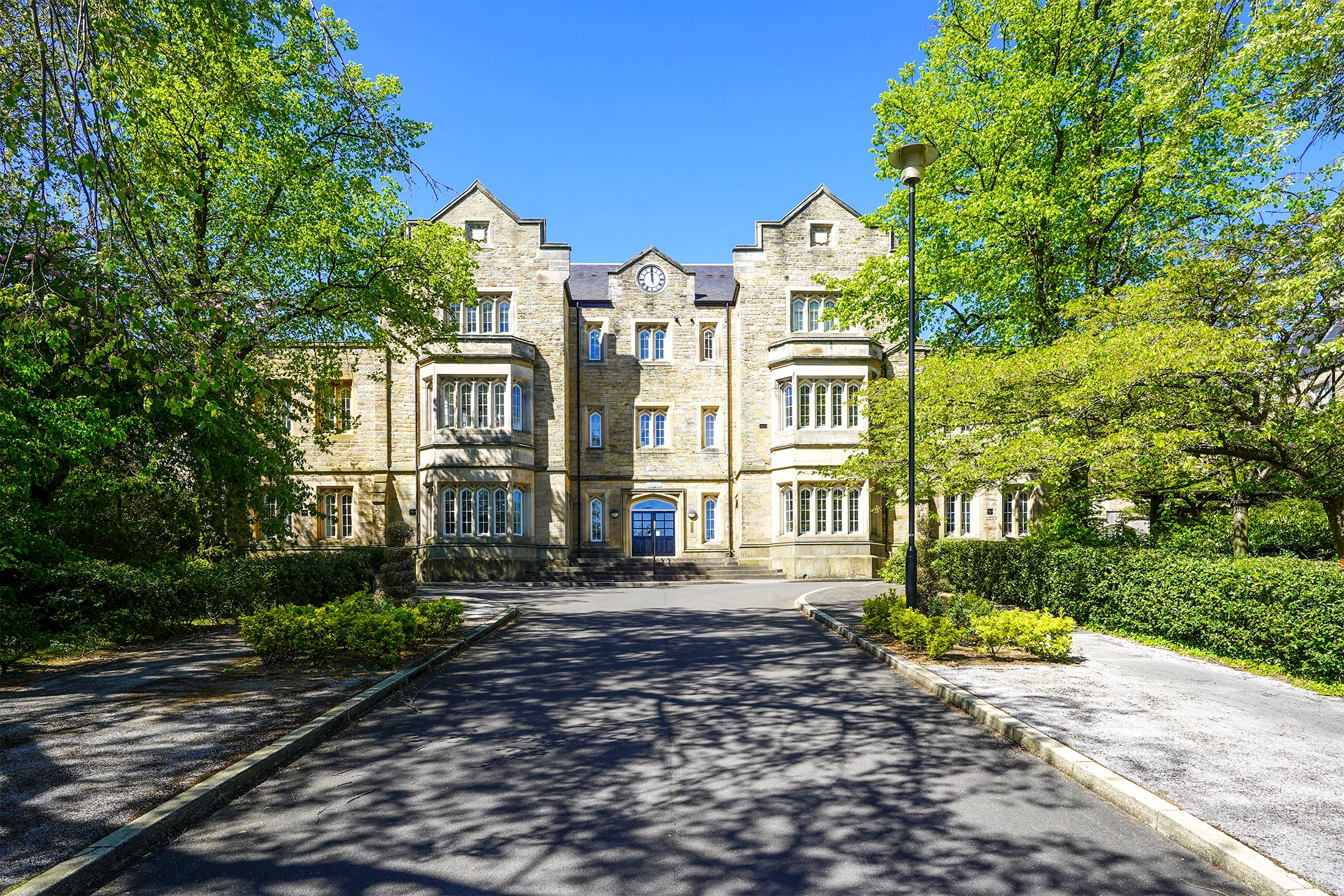 Stone building with clock tower, blue entrance door, multiple windows, and driveway lined with green hedges and trees