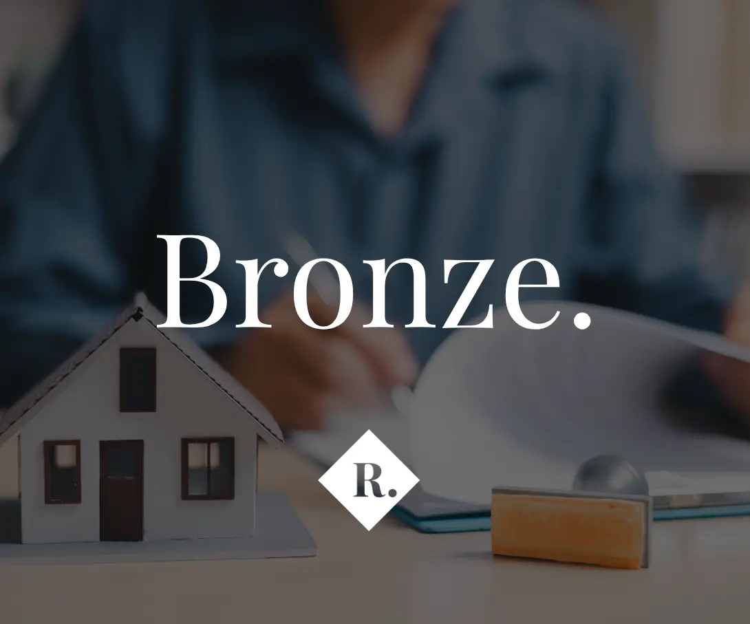 Miniature house model and wooden stamp on desk with blurred figures in background. Text overlay reads "Bronze." with "R." logo