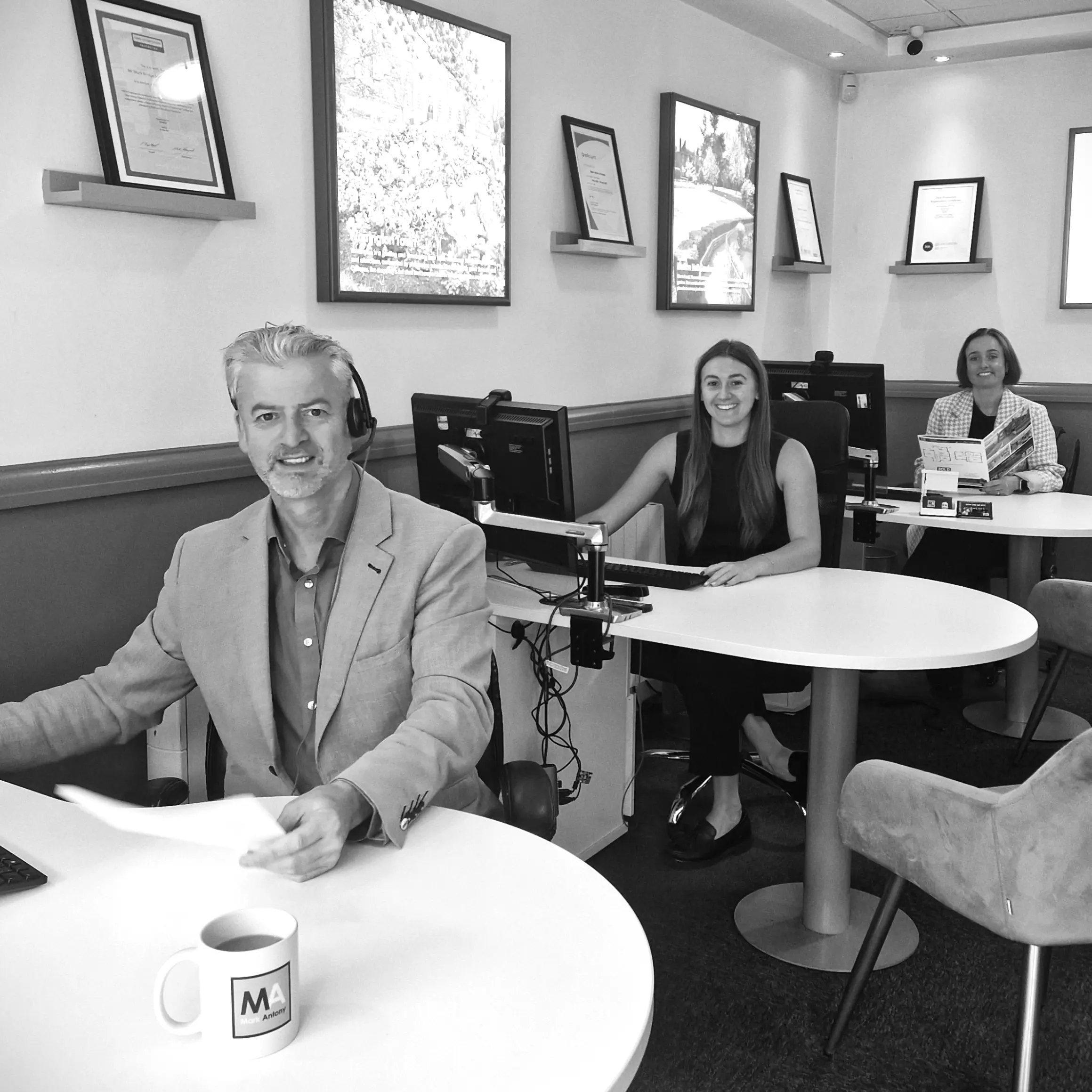 Three people in an office with computers and framed photographs on walls. Man in foreground wearing headphones and blazer at round table with mug.