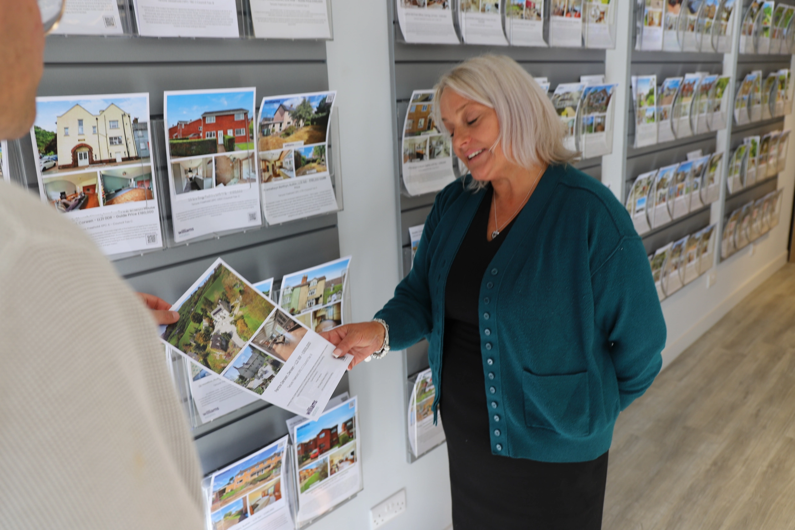 Woman in teal cardigan holding property brochure in front of estate agency window display with property listings