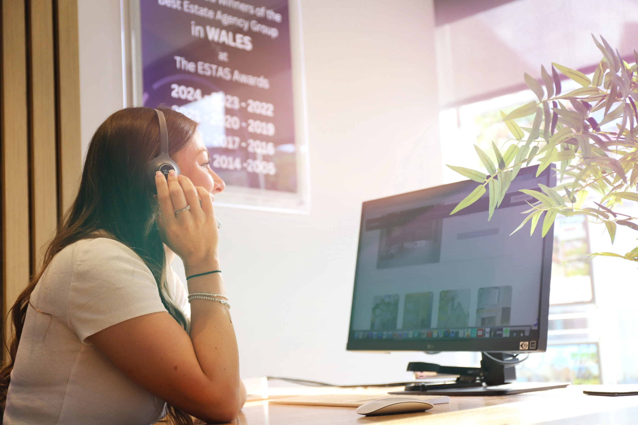 Person at desk with computer monitor displaying a property image, with a purple sign mentioning ESTAS Awards visible on the wall behind