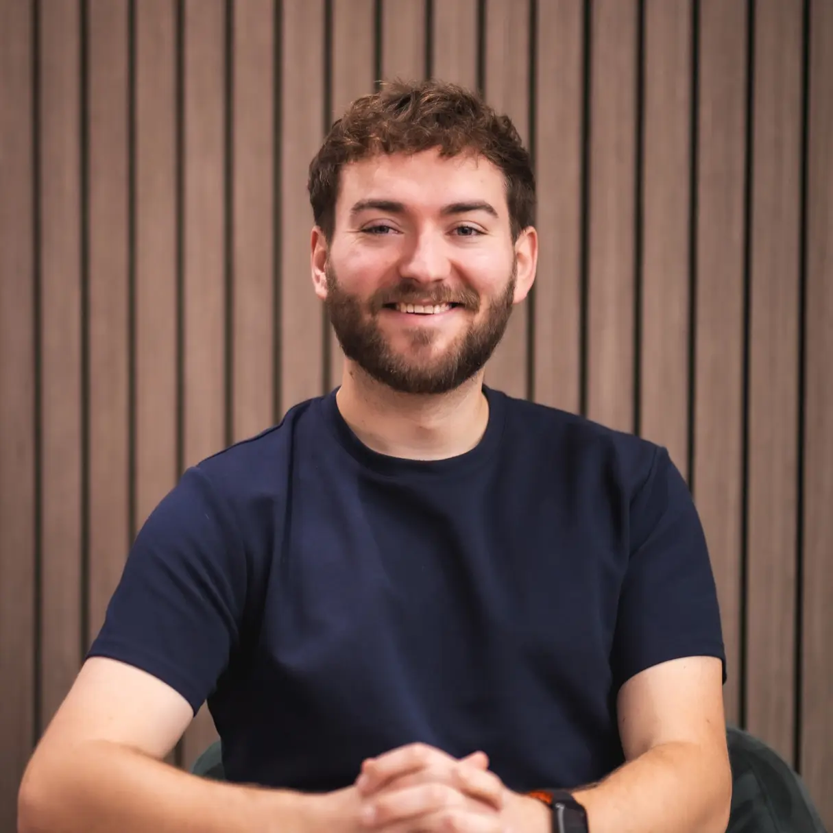 Man with dark hair and beard wearing navy t-shirt, smiling at camera against striped brown wall