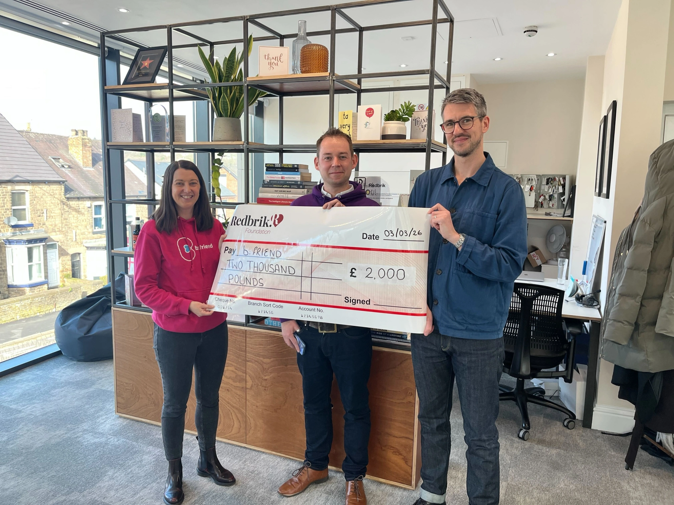 Three people holding a large cheque for £2,000 from Redbrik to b.FRIEND, dated 03/03/26, in an office with shelving and windows