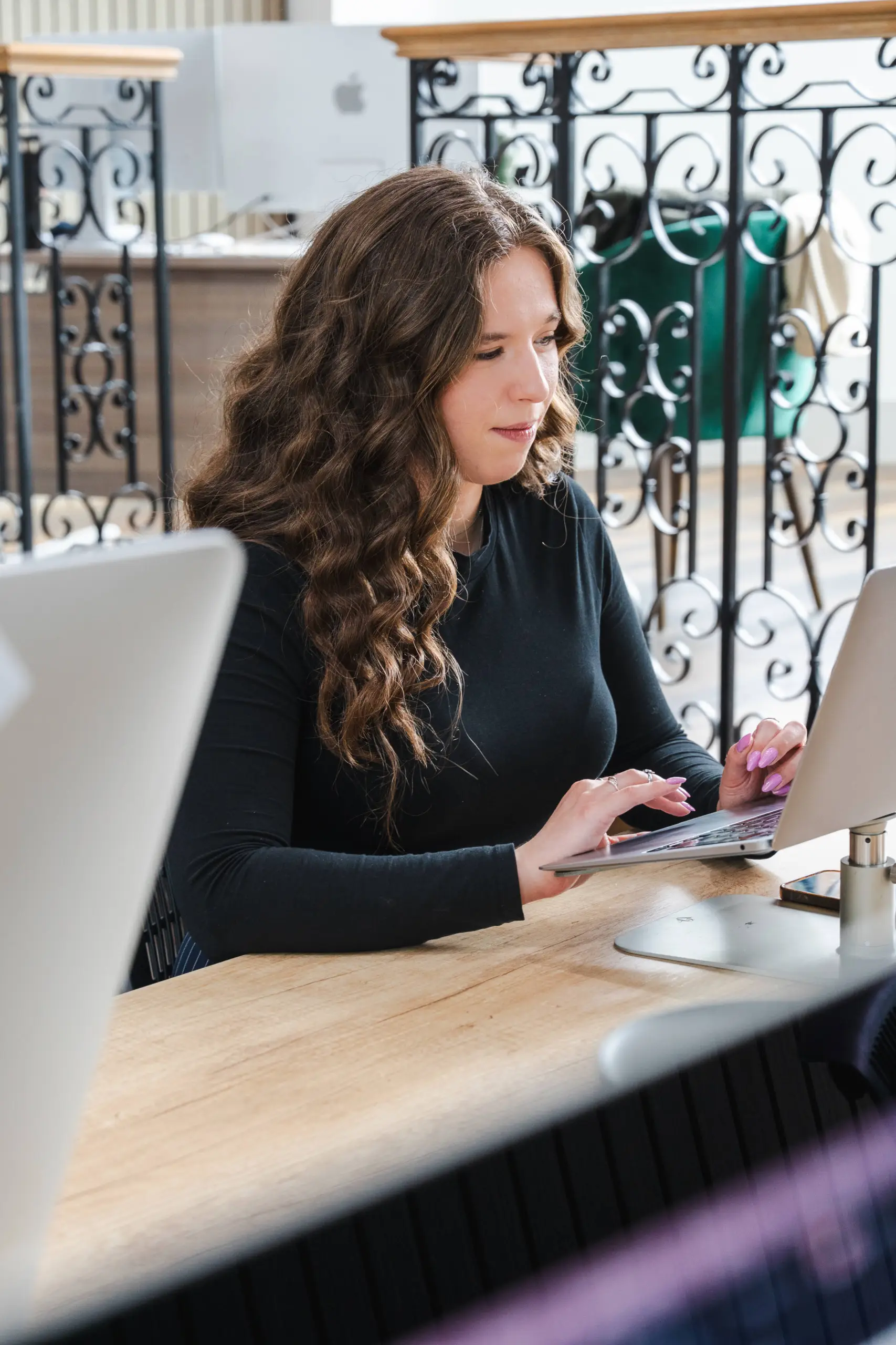 Woman with long brown wavy hair wearing black long-sleeved top, sitting at wooden desk with laptop and computer mouse, ornate black and white metal railings visible in background