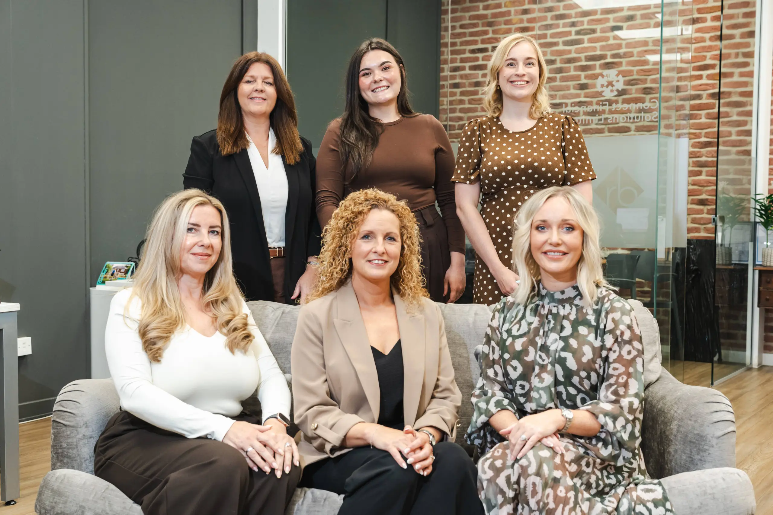 Six women in an office with exposed brick wall, three seated on a grey sofa and three standing behind