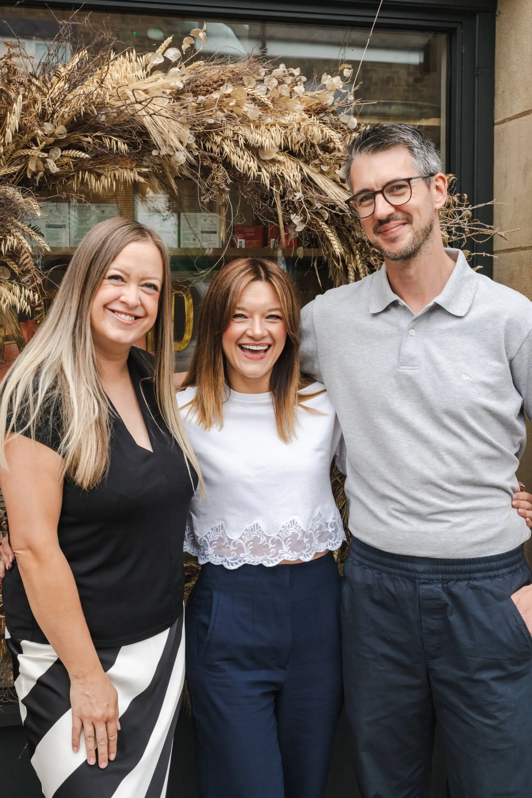 Three people standing outside a storefront with dried floral decoration above