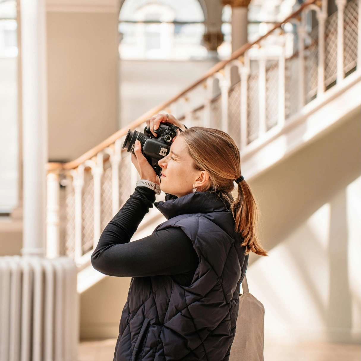 Person holding a camera to their face, standing in a stairwell with white railings and wooden handrails