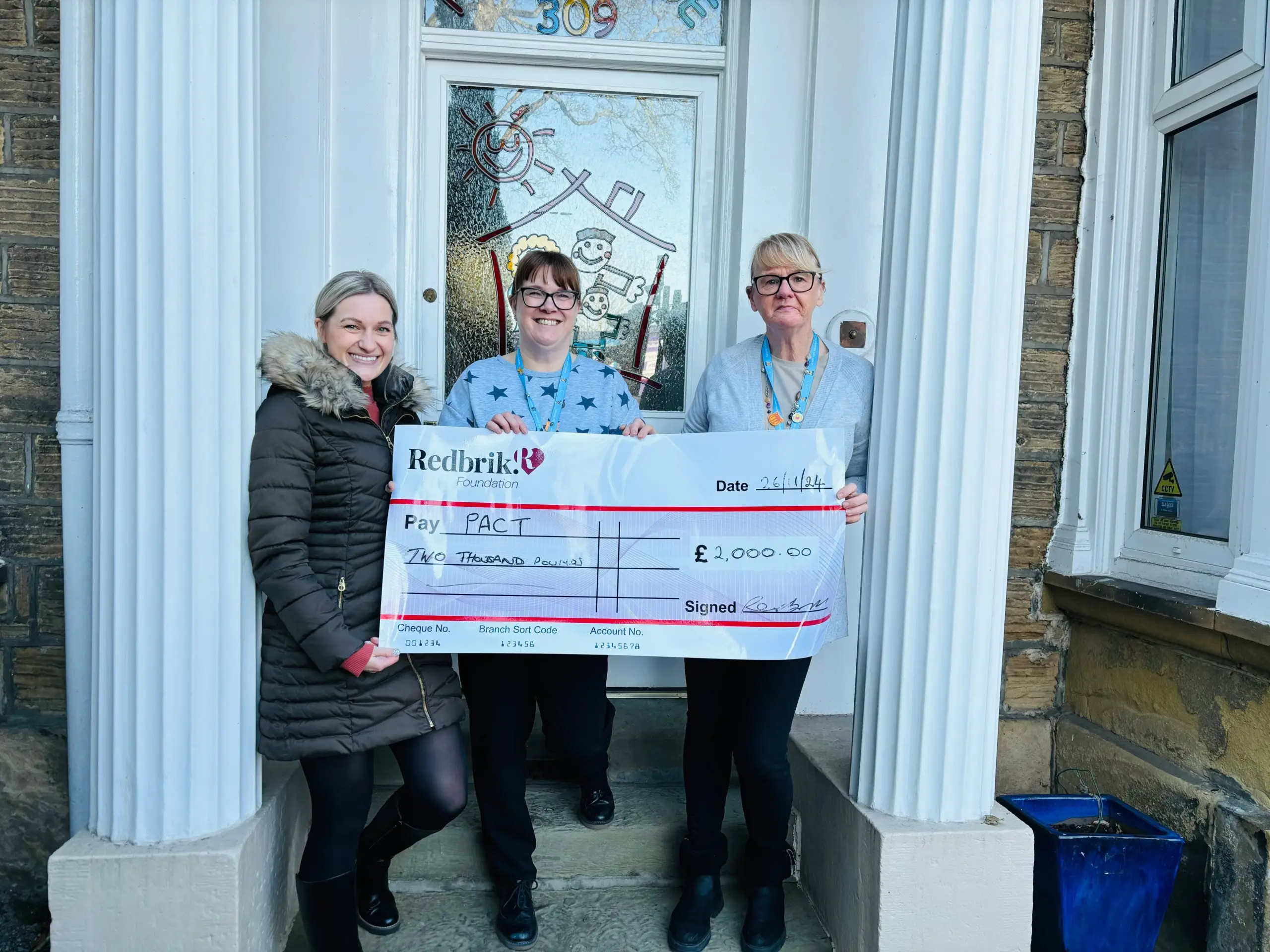 Three people holding a large cheque for £2,000 from Redbrick Foundation to PACT, dated 26/11/24, standing in a doorway numbered 309