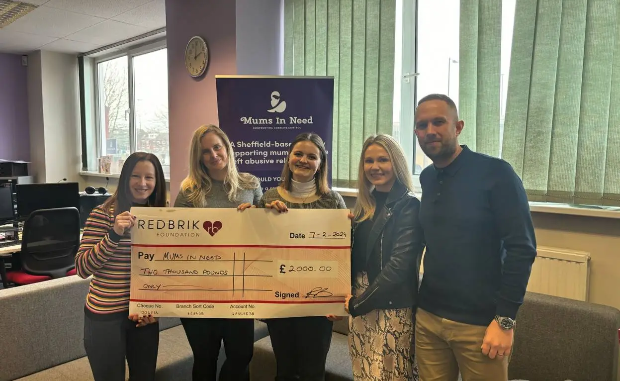 Five people holding a large cheque for £2,000 made payable to Mums in Need, dated 7-2-2024, in an office with a Mums in Need banner visible behind them