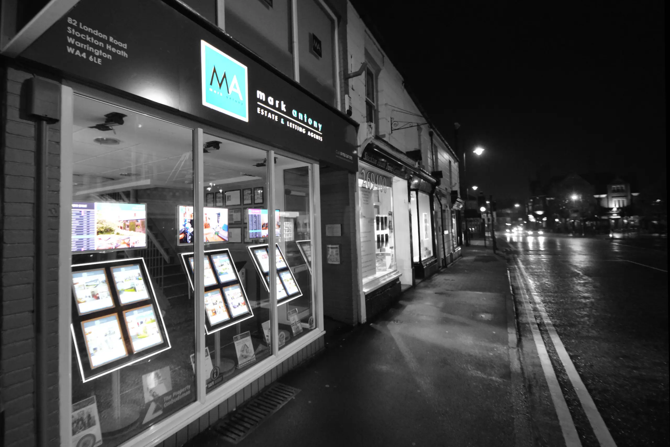 Mark Unleash estate agent storefront at night, with illuminated window displays showing property listings and a cyan logo sign above
