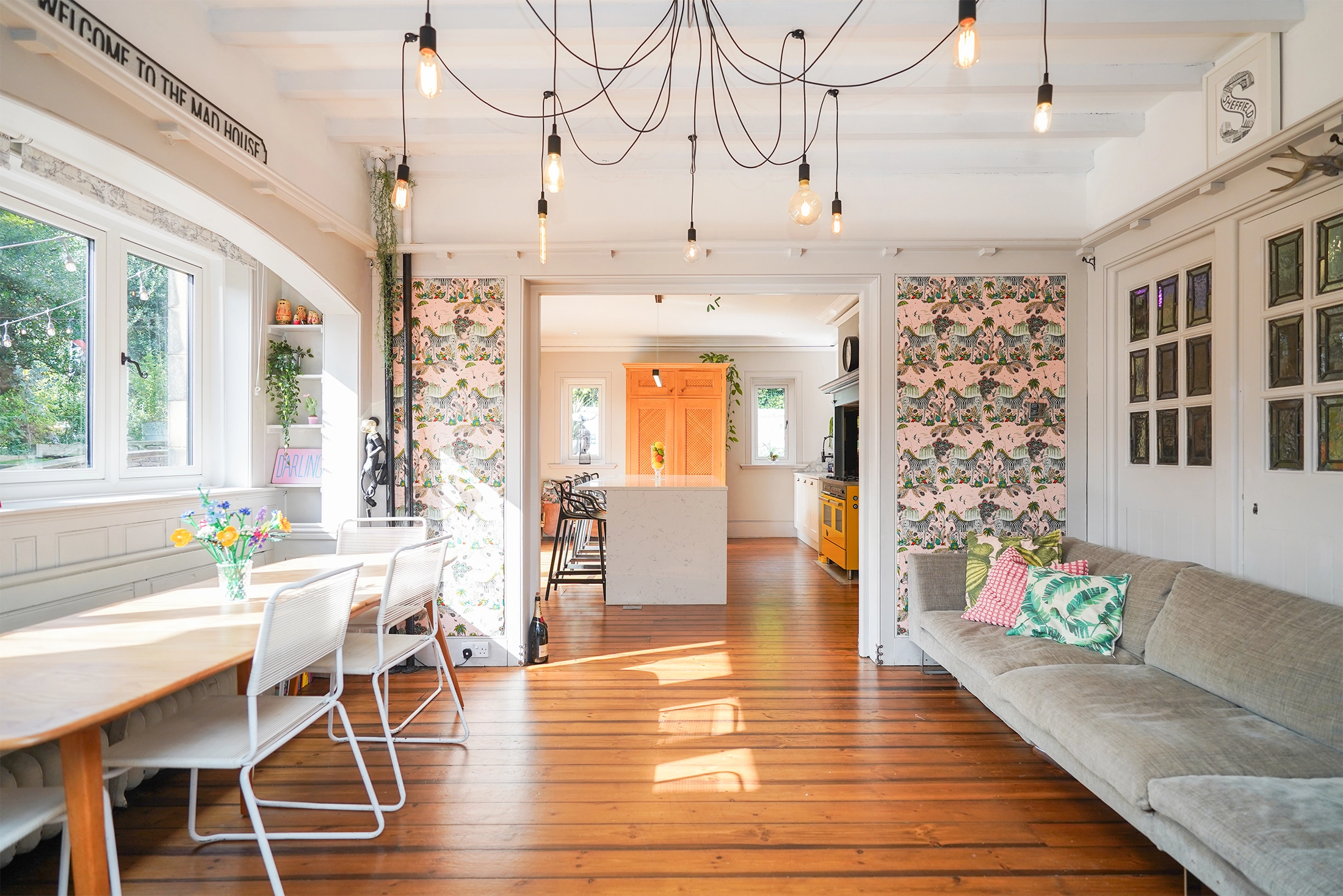 Open-plan kitchen and living room in Edwardian house with wooden flooring, pendant lights, floral wallpaper, orange front door visible through archway