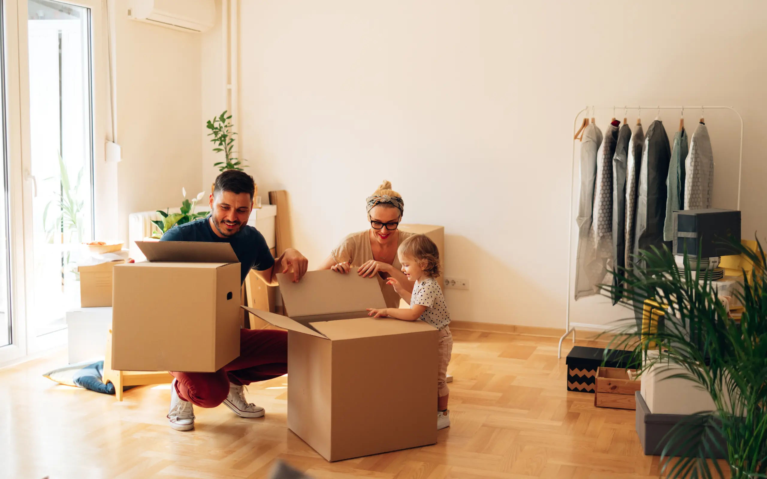 Three people unpacking cardboard boxes in a bright, empty room with wooden flooring, plants, and a clothing rail