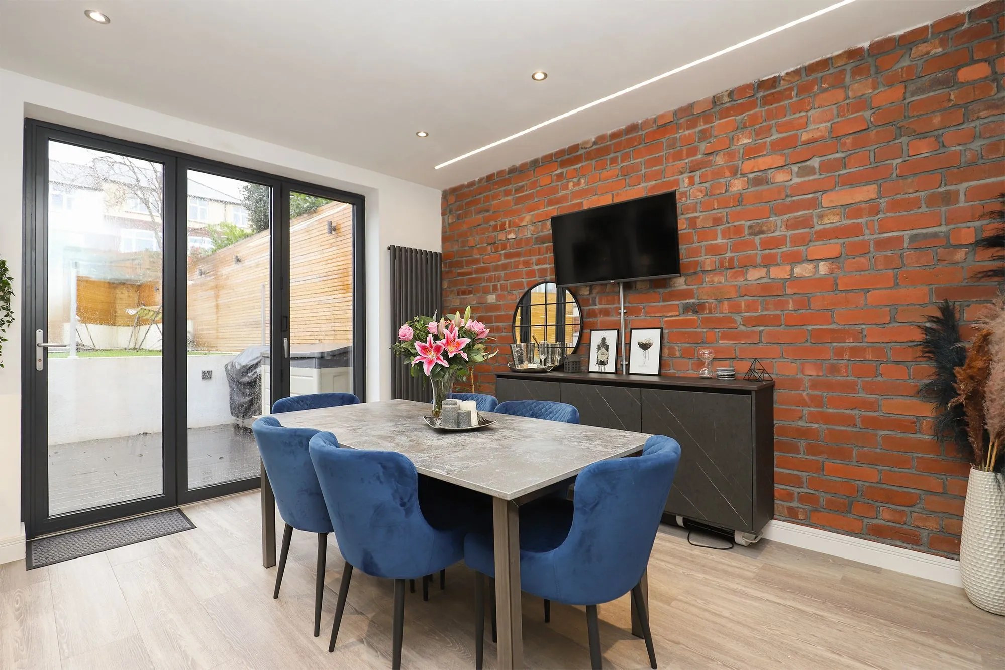 Modern dining room with exposed red brick wall, grey stone dining table, four blue velvet chairs, wall-mounted TV, black sideboard with flowers, and glass doors opening to garden