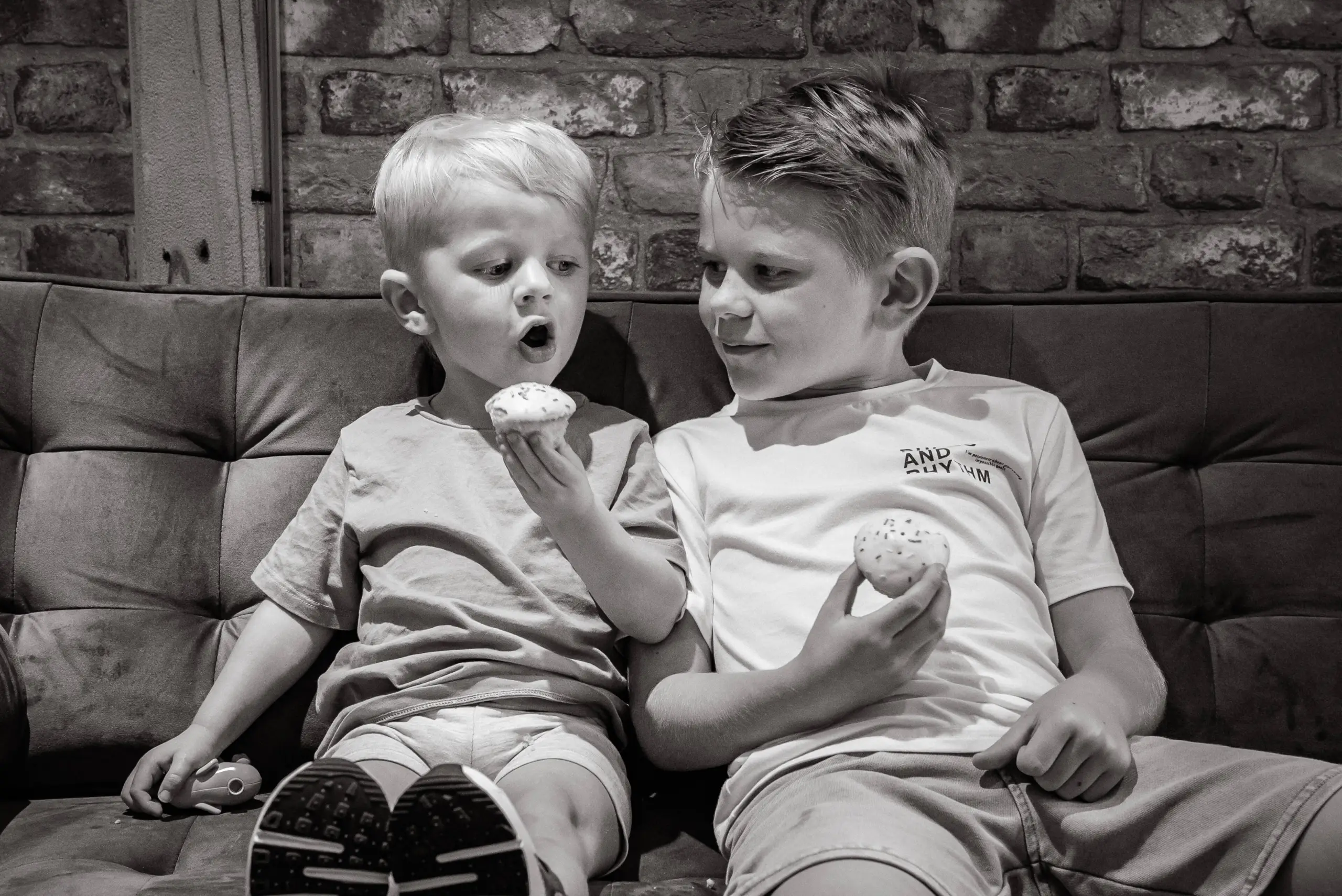 Two young children sitting on a sofa eating cupcakes, with a brick wall behind them