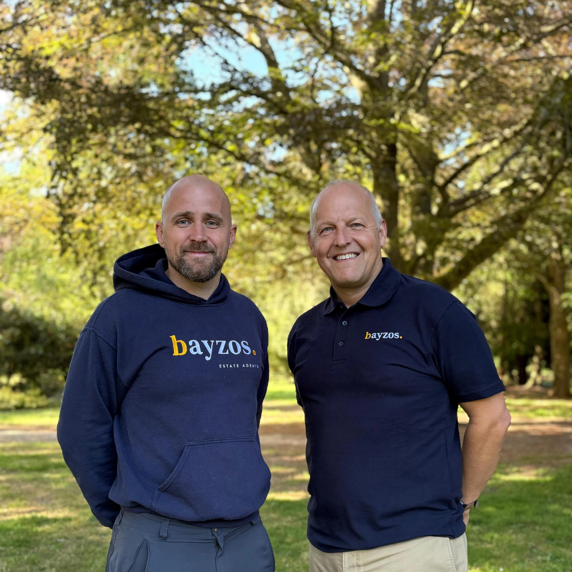 Two men standing outdoors under trees, both wearing navy Bayzos branded clothing