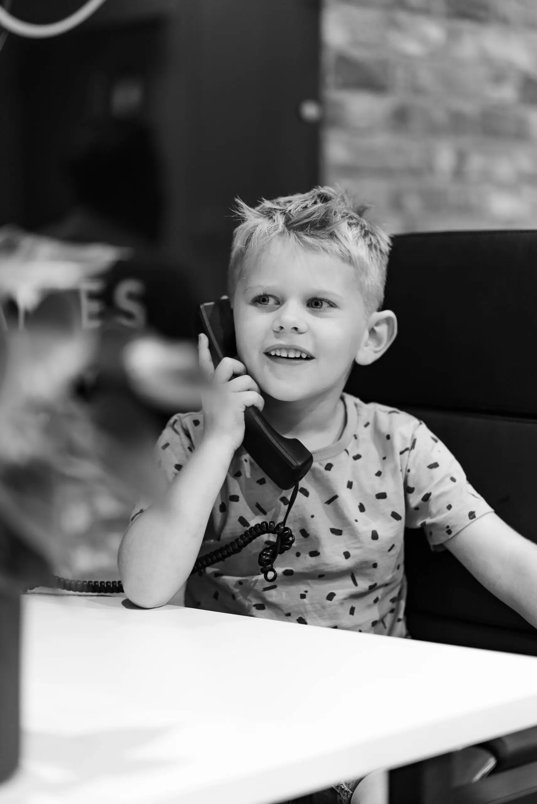 Young child holding a telephone handset to their ear, smiling, seated at a desk in black and white
